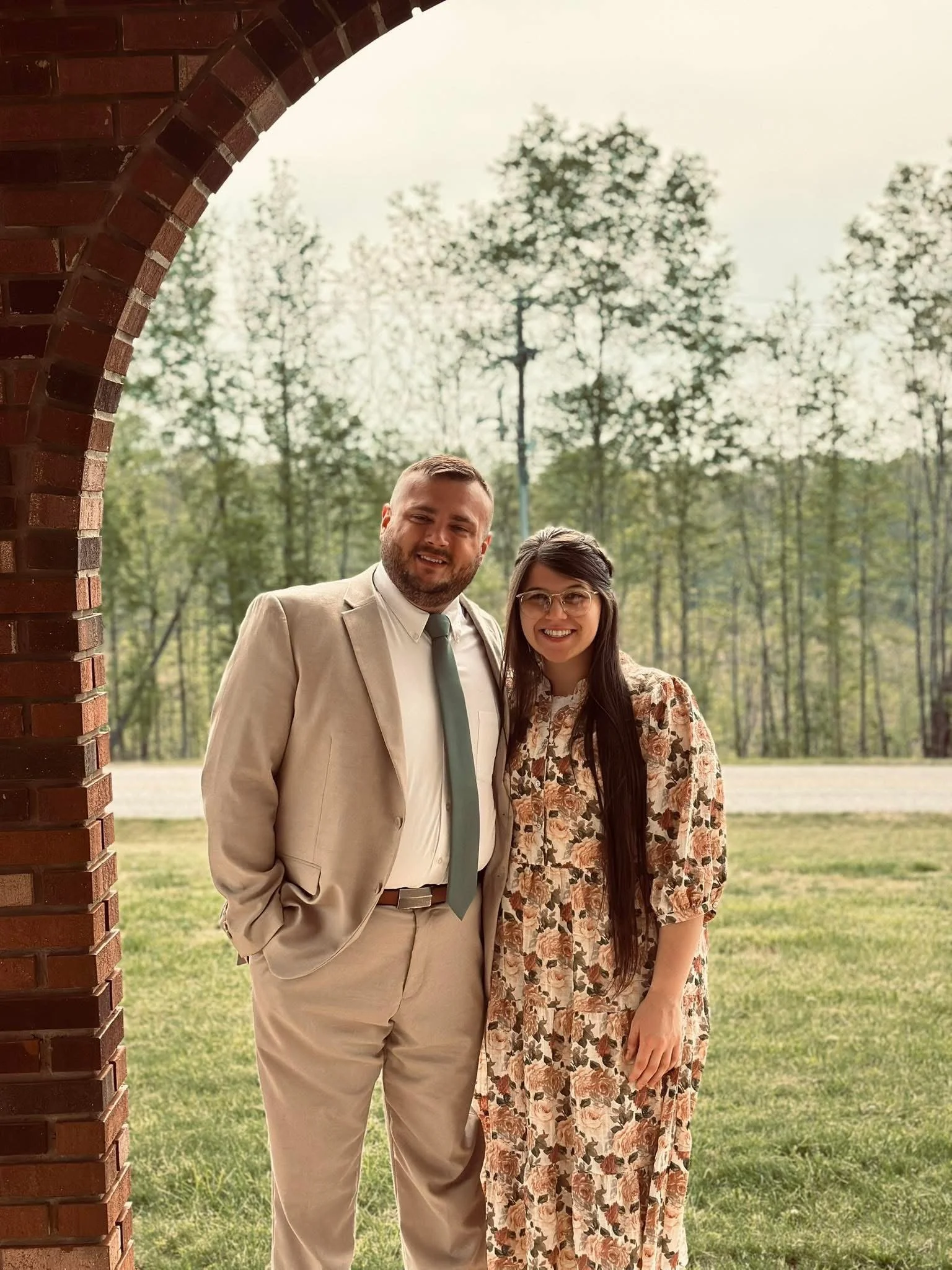 A man and a woman standing outside under an archway made of bricks, with trees and a grassy area in the background, smiling at the camera.