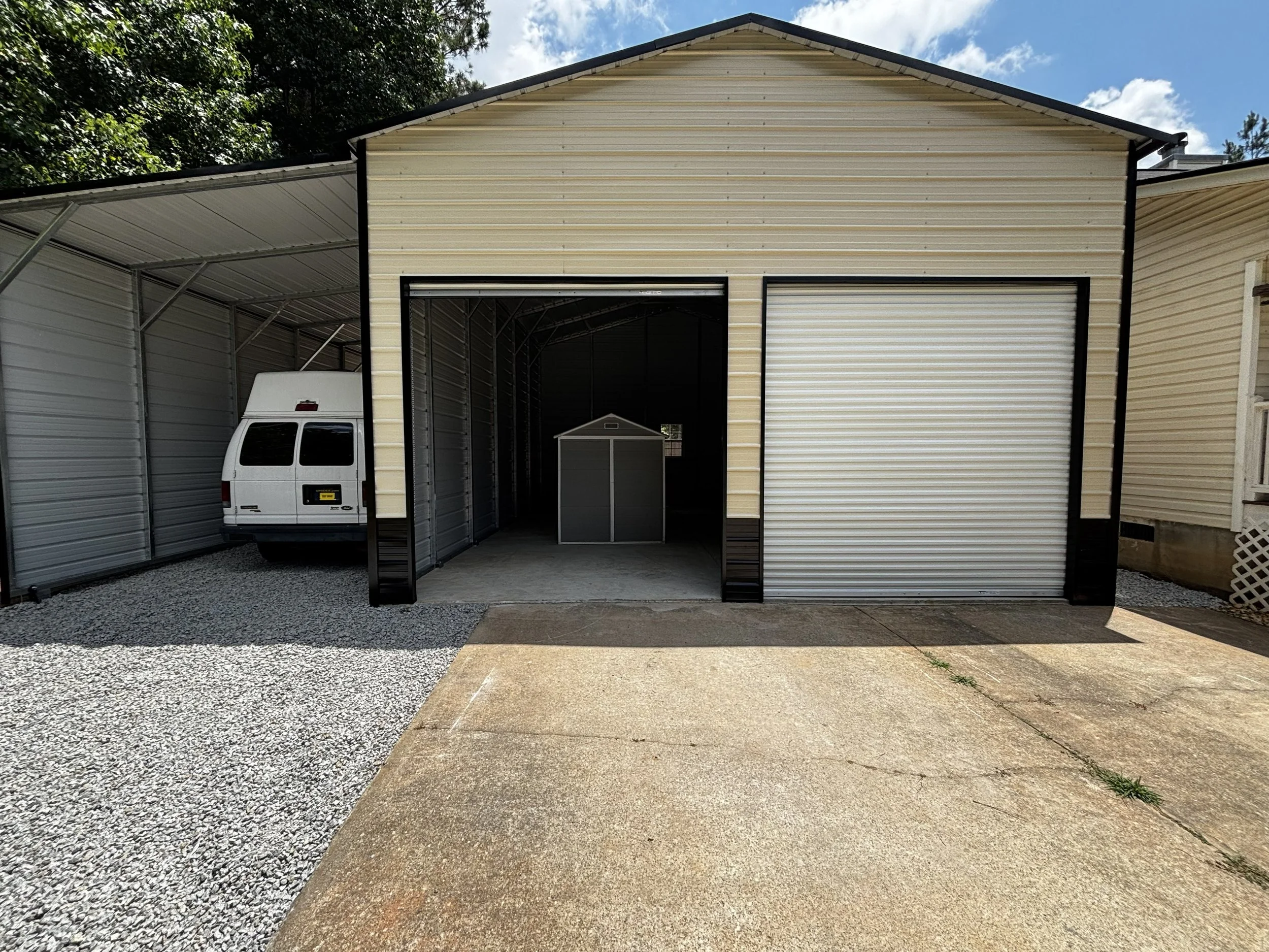A storage shed with metal siding, open garage door, and a white van parked underneath a metal carport. The shed is situated on a gravel driveway with a paved section in front, surrounded by trees and blue sky with some clouds.