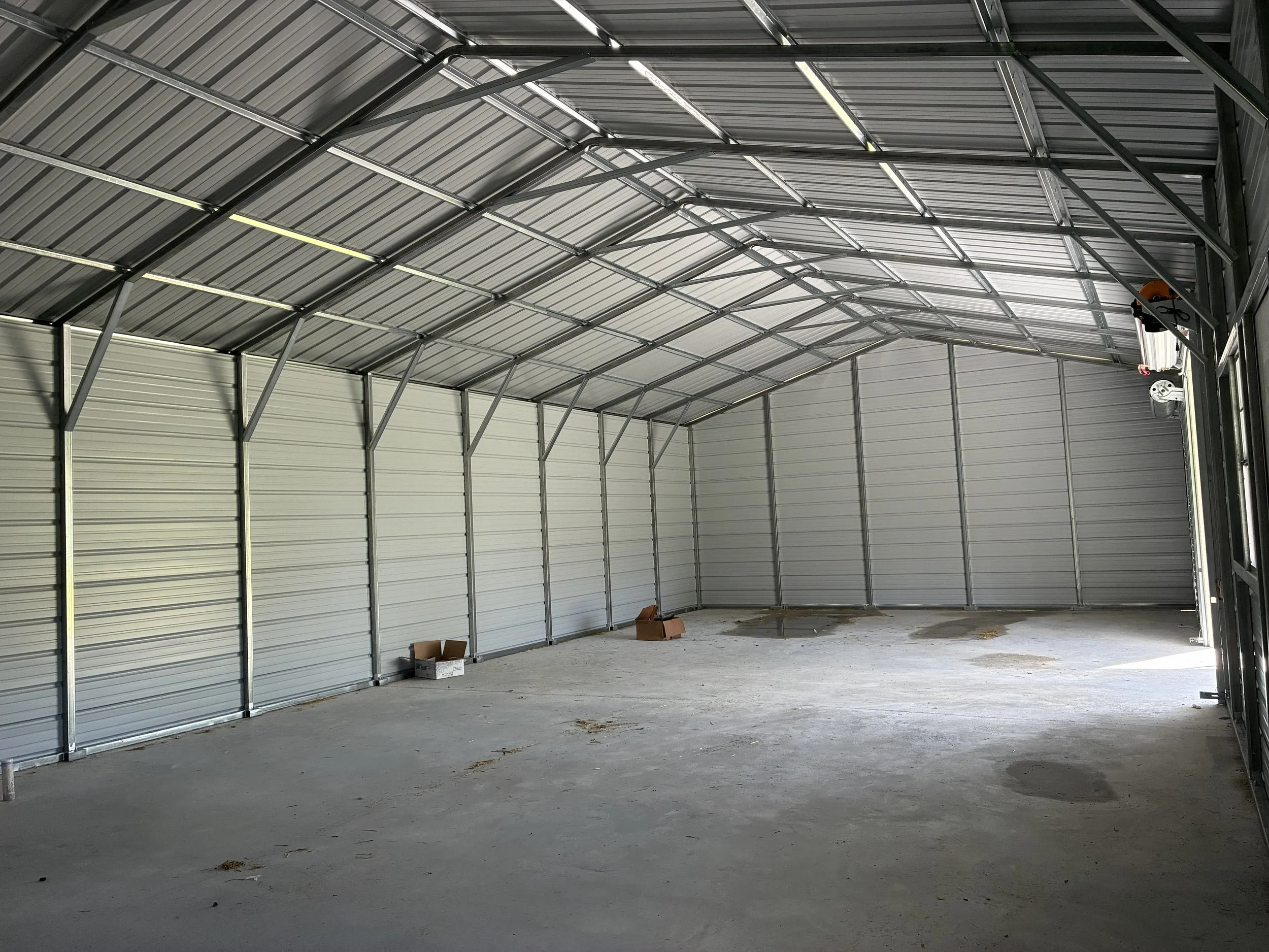 Interior of an empty, unfinished garage with metal walls, a high arched ceiling, a concrete floor, and some cardboard boxes near the back wall.