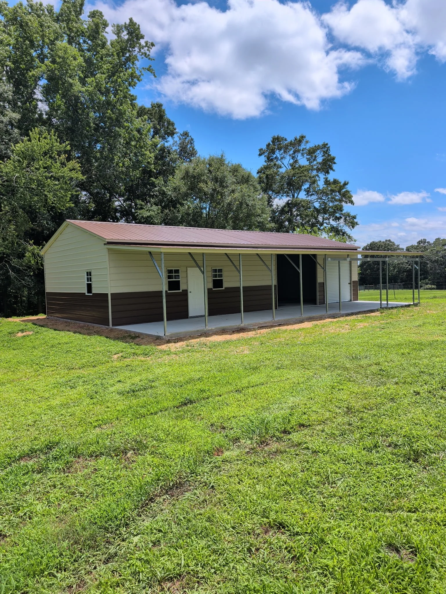 A small beige garage with a maroon metal roof and open framing in the front, situated on a grassy lawn with trees and a partly cloudy blue sky in the background.