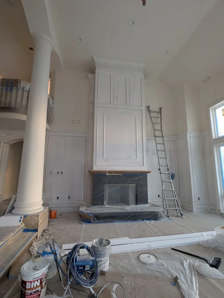 Interior of a room under construction or renovation with a white ceiling, large windows, a decorative built-in cabinet or fireplace with a dark stone finish, a ladder, and construction supplies on the floor.
