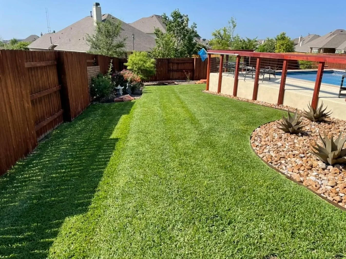 Well-maintained backyard lawn with a wooden privacy fence on the left and a deck with a railing on the right, featuring a swimming pool and lounge chairs, with various plants and trees in the background.