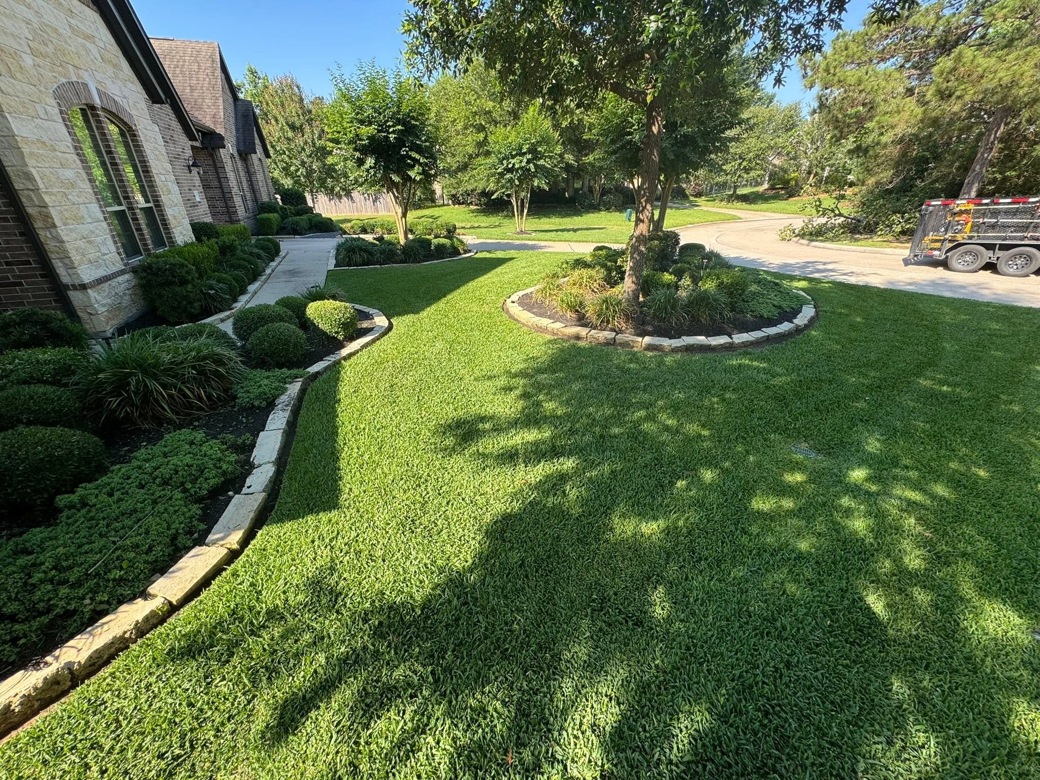 A residential front yard with a landscaped garden, green grass, and several trees. There is a sidewalk and a driveway with a trailer parked on it. The house has brick walls and multiple windows.