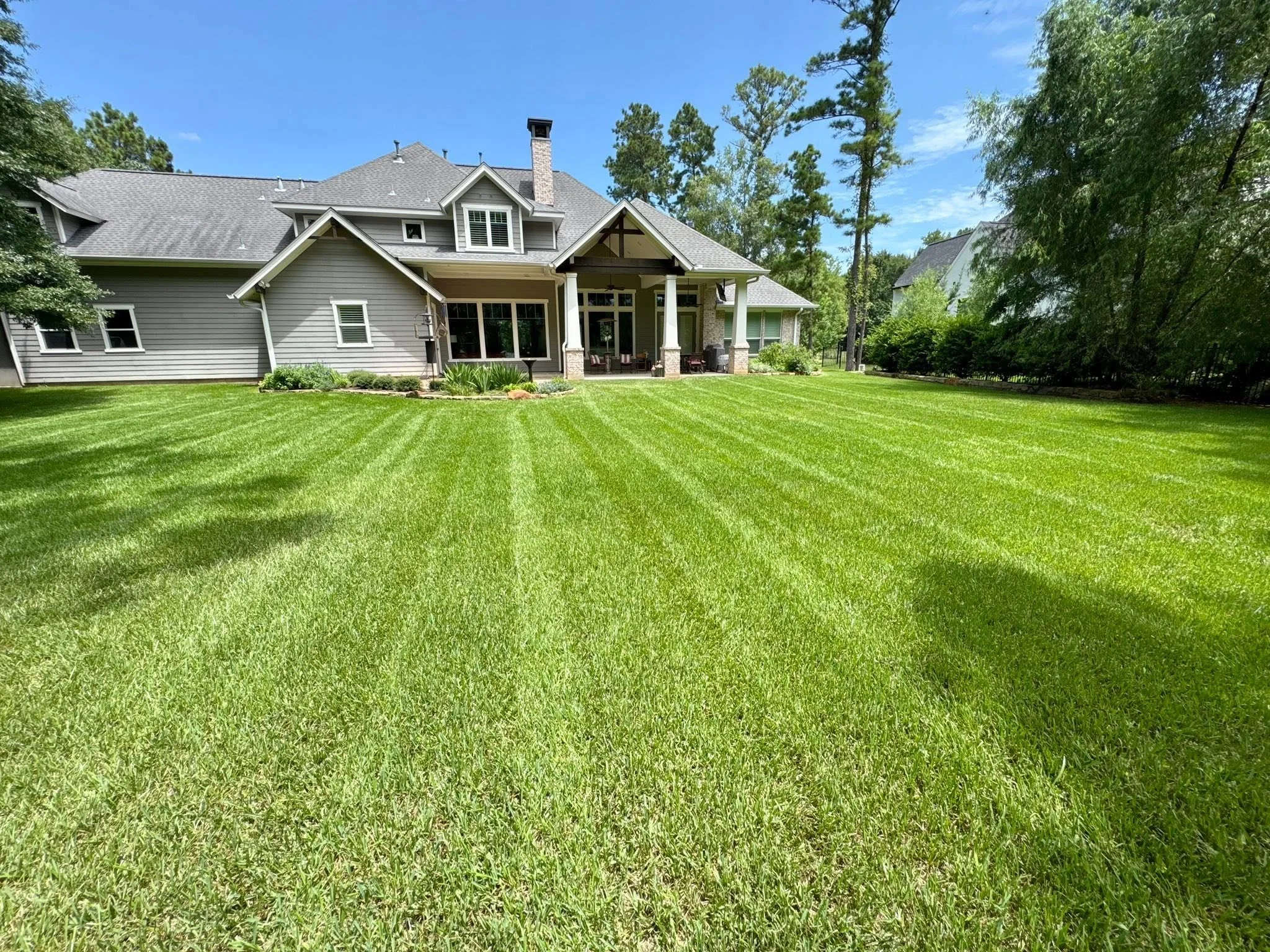 A large house with a gray exterior, multiple gabled roofs, white window shutters, and a covered porch with outdoor furniture. The house is surrounded by a well-maintained, lush green lawn and tall trees under a clear blue sky.