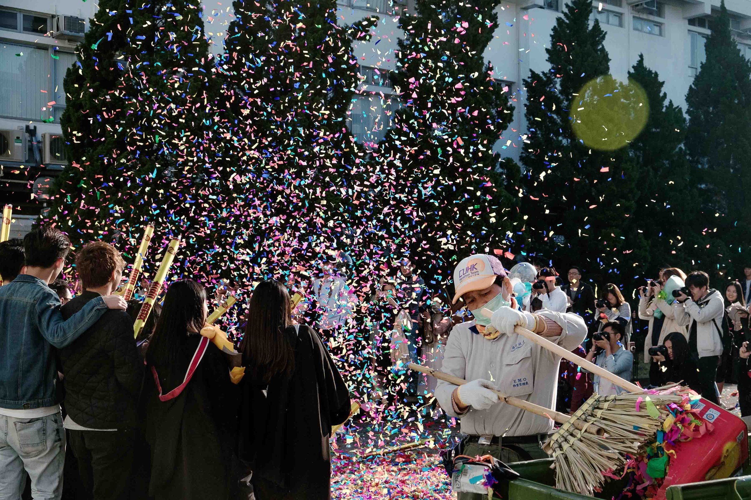 People celebrating in an outdoor event with confetti, some wearing masks, with buildings and trees in the background.