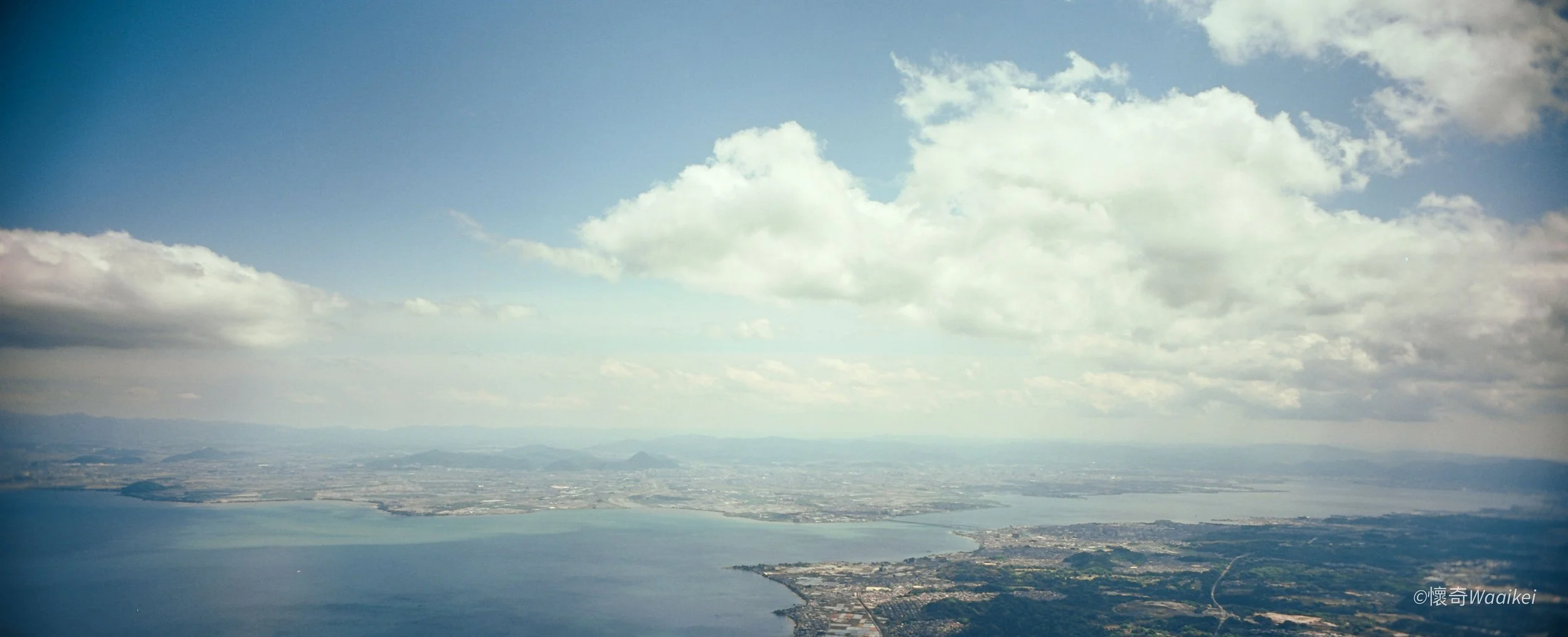 Aerial view of a coastal city with a large body of water, land, and a cloudy sky.