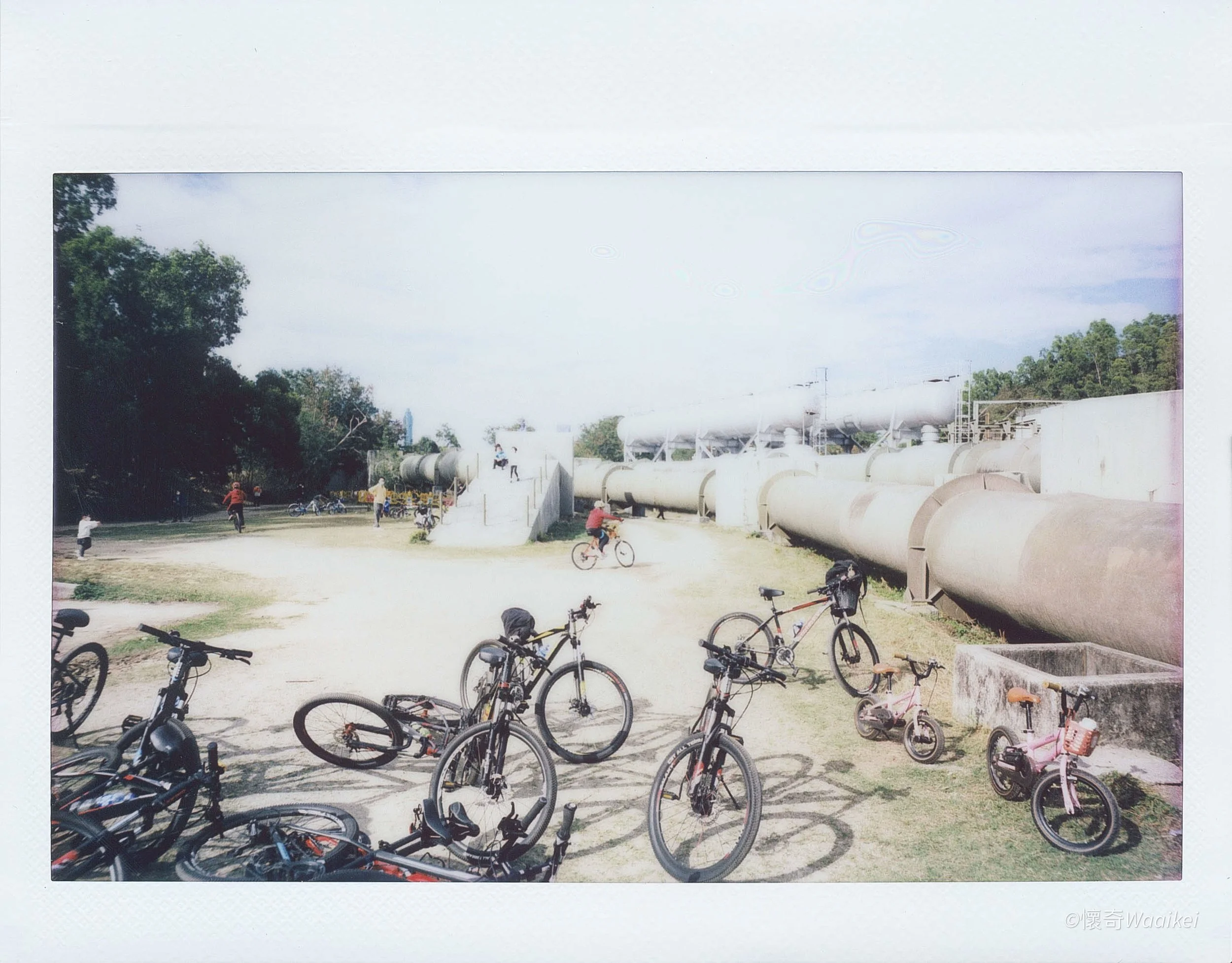 A park area with multiple bicycles parked in the foreground, children and adults playing and riding bikes, large industrial pipes on the right, a white wall, and trees with a cloudy sky in the background.