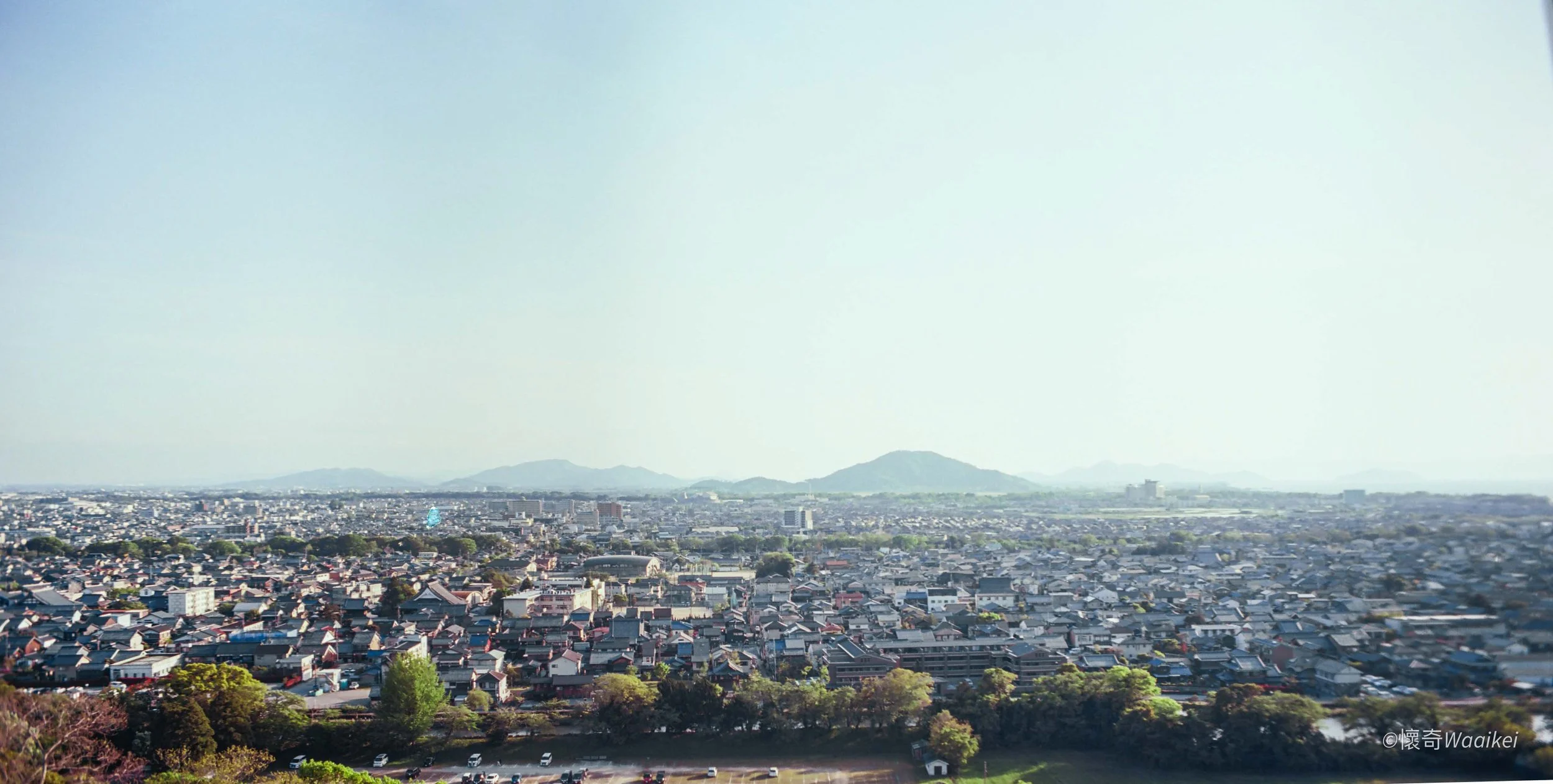 A panoramic view of a city with numerous residential and commercial buildings, distant mountains under a clear sky, and some trees in the foreground.