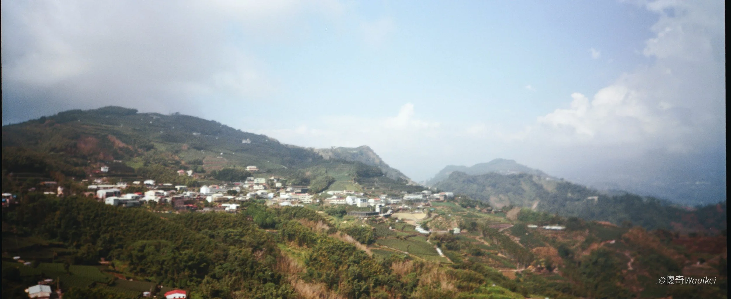 Hilly landscape with green trees and mountains, scattered houses, and a partly cloudy sky.