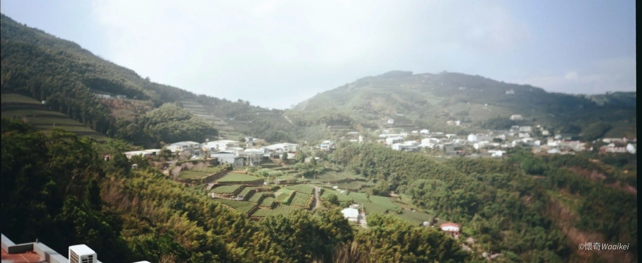 A panoramic view of a hillside town with numerous white buildings, surrounded by lush green trees and terraces on the mountainside, under a partly cloudy sky.