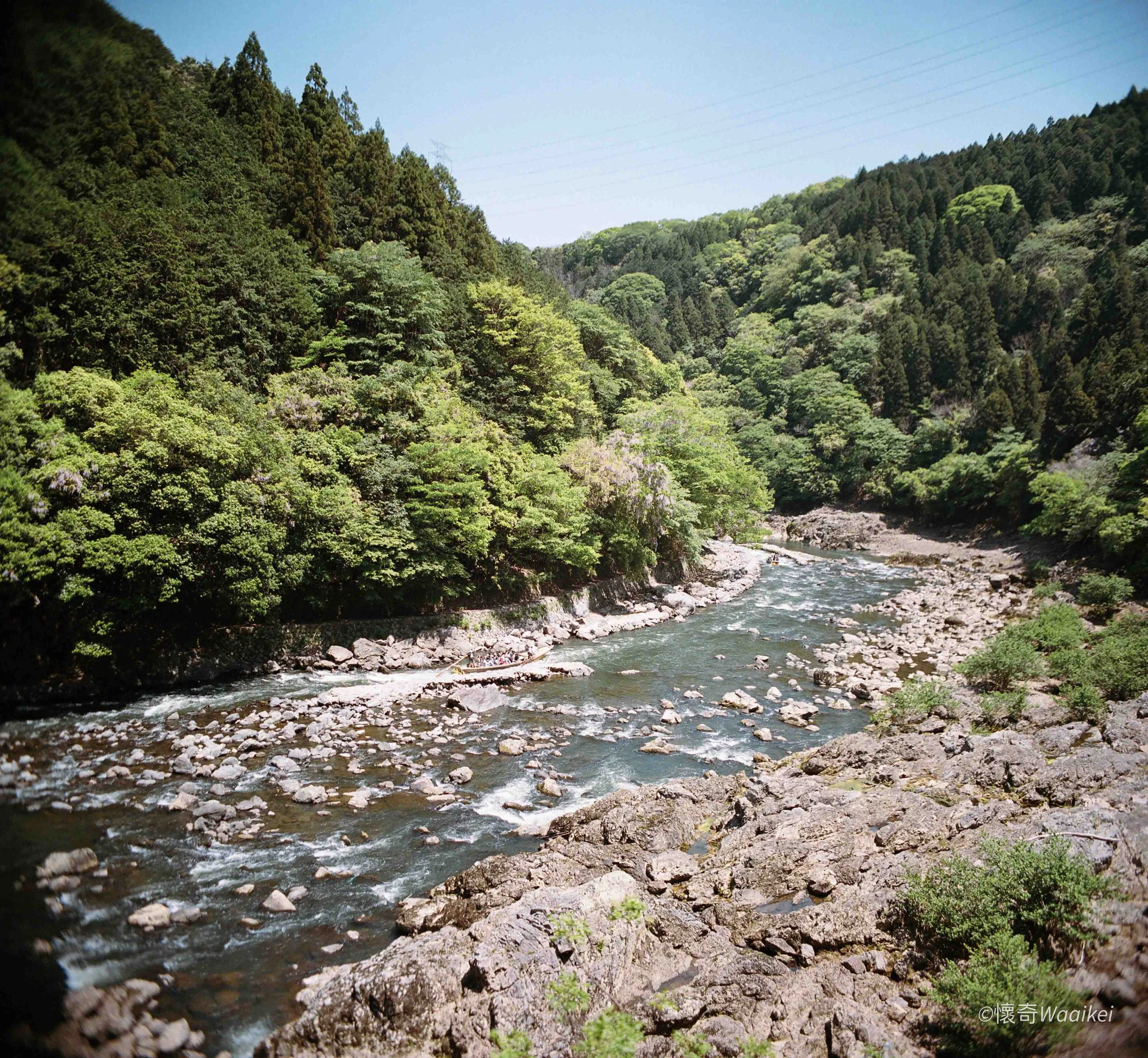 A scenic view of a fast-flowing river with rocks along the banks surrounded by dense green trees and mountains under a clear blue sky.