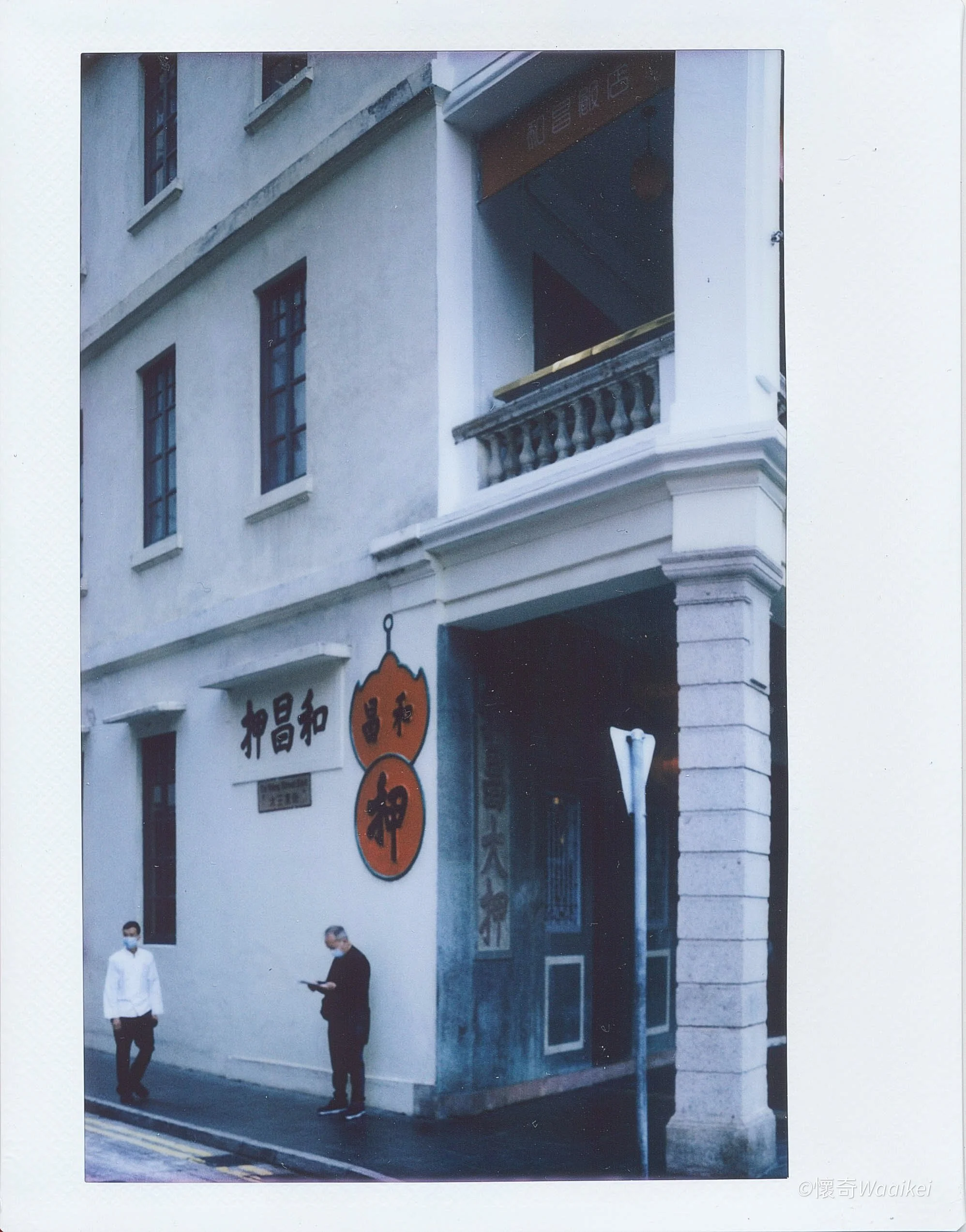 Photograph of a building with Japanese signboards and two people standing on the sidewalk; one person is in a white coat and face mask, the other in dark clothing and holding a phone.