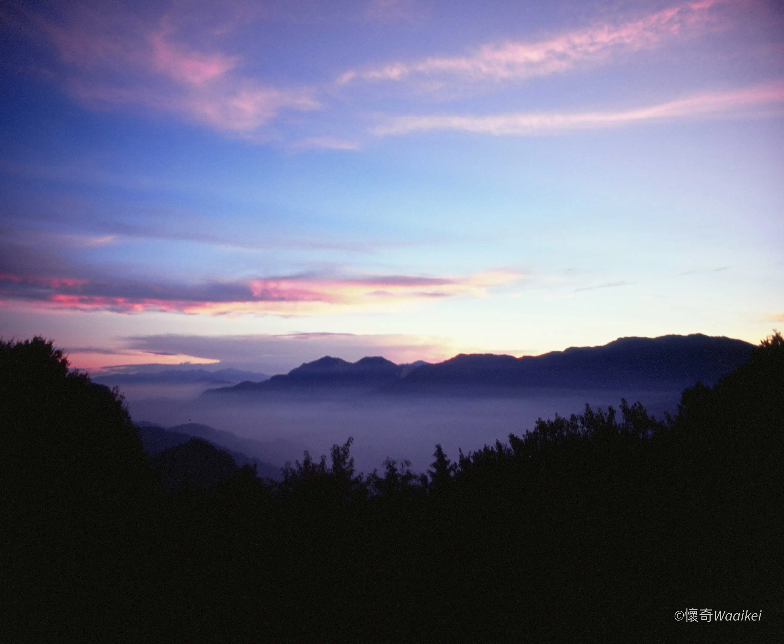 A landscape view of mountains at dawn with a colorful sky featuring pink and purple clouds, and dark silhouette of trees in the foreground.
