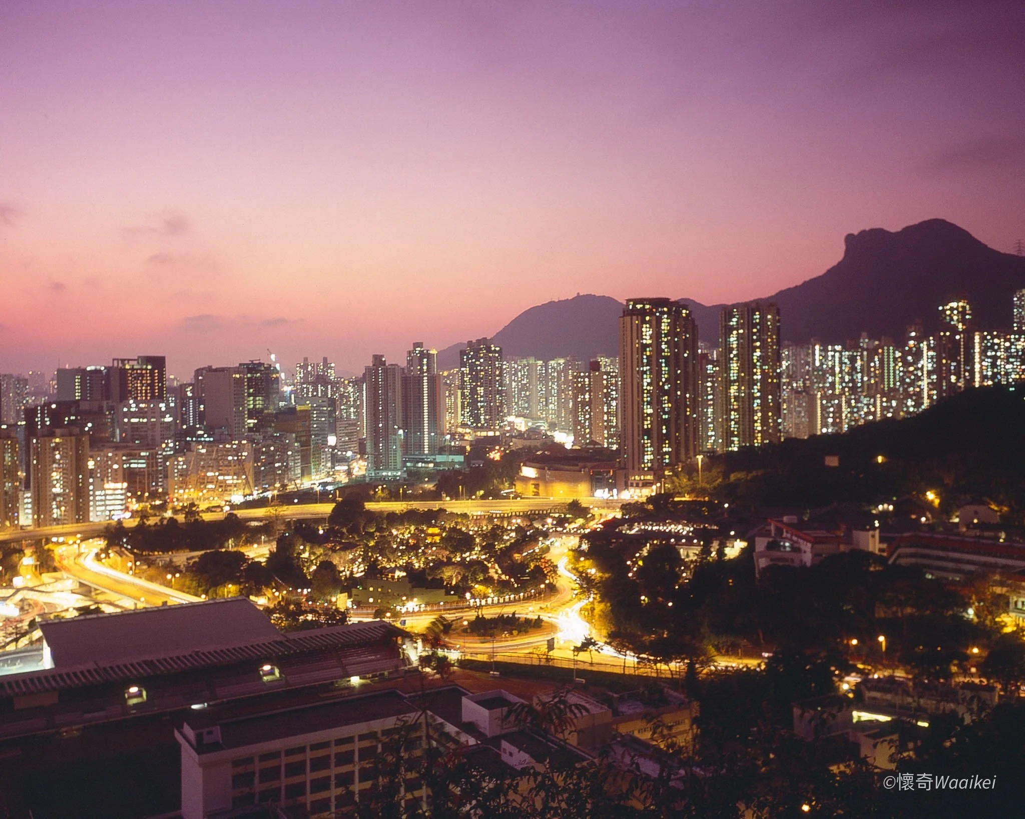 City skyline at dusk with tall buildings, mountain background, and pink-purple sky.