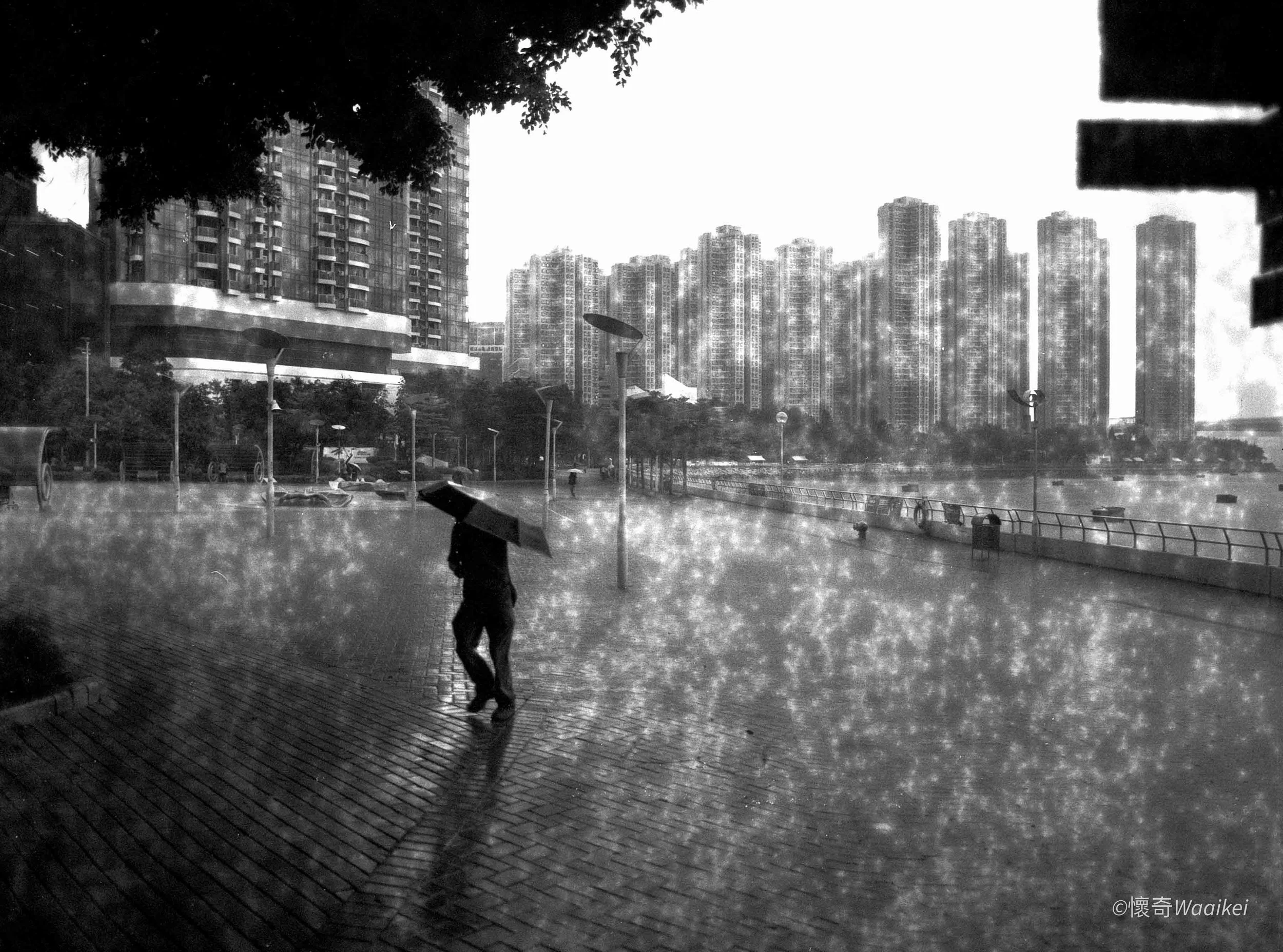 A person walking with an umbrella on a rainy, wet sidewalk in an urban cityscape with tall buildings in the background.