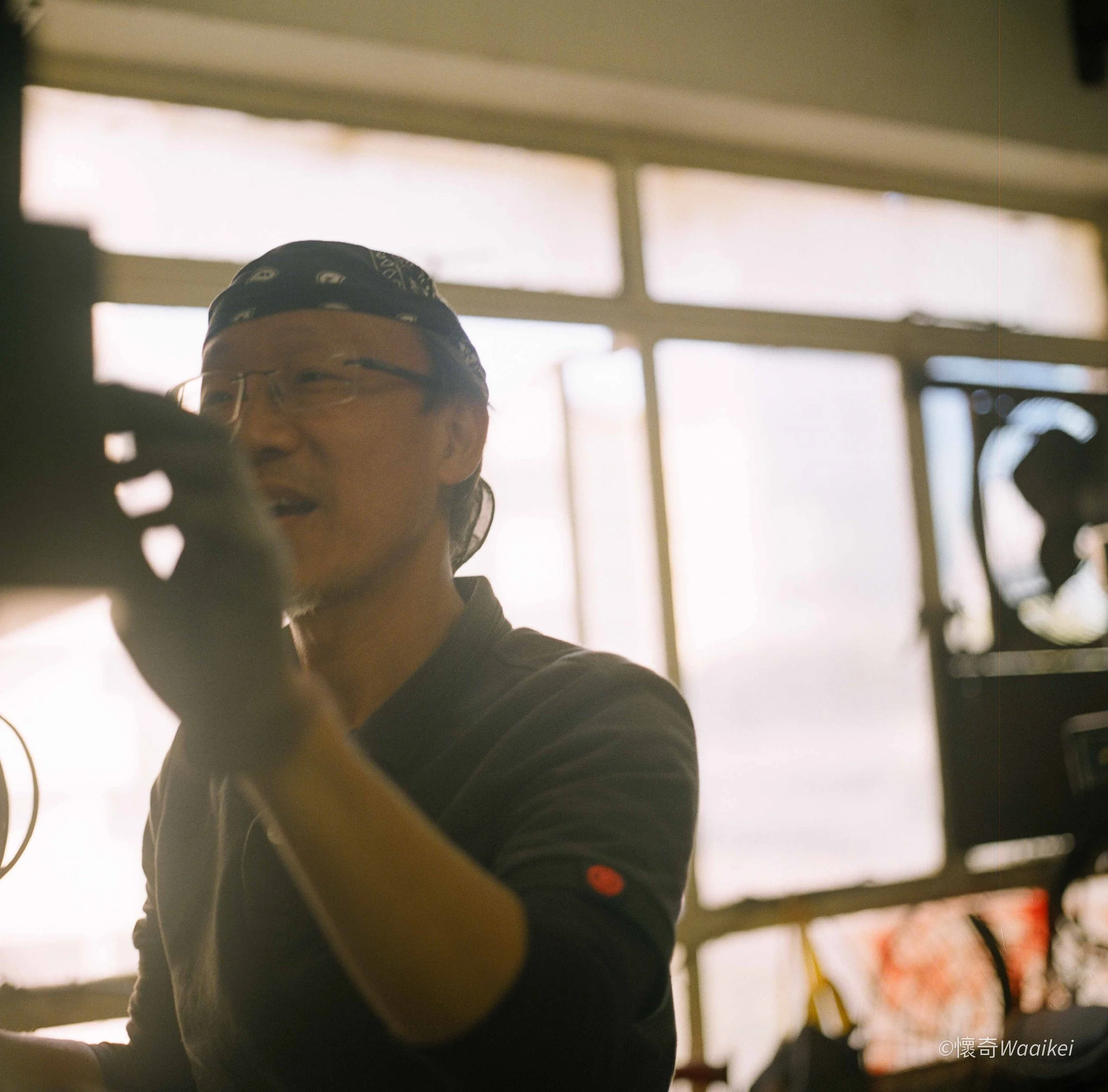 A person wearing glasses, a black bandana, and a black shirt smiling and looking at the camera. The background shows blurred windows and bright light.
