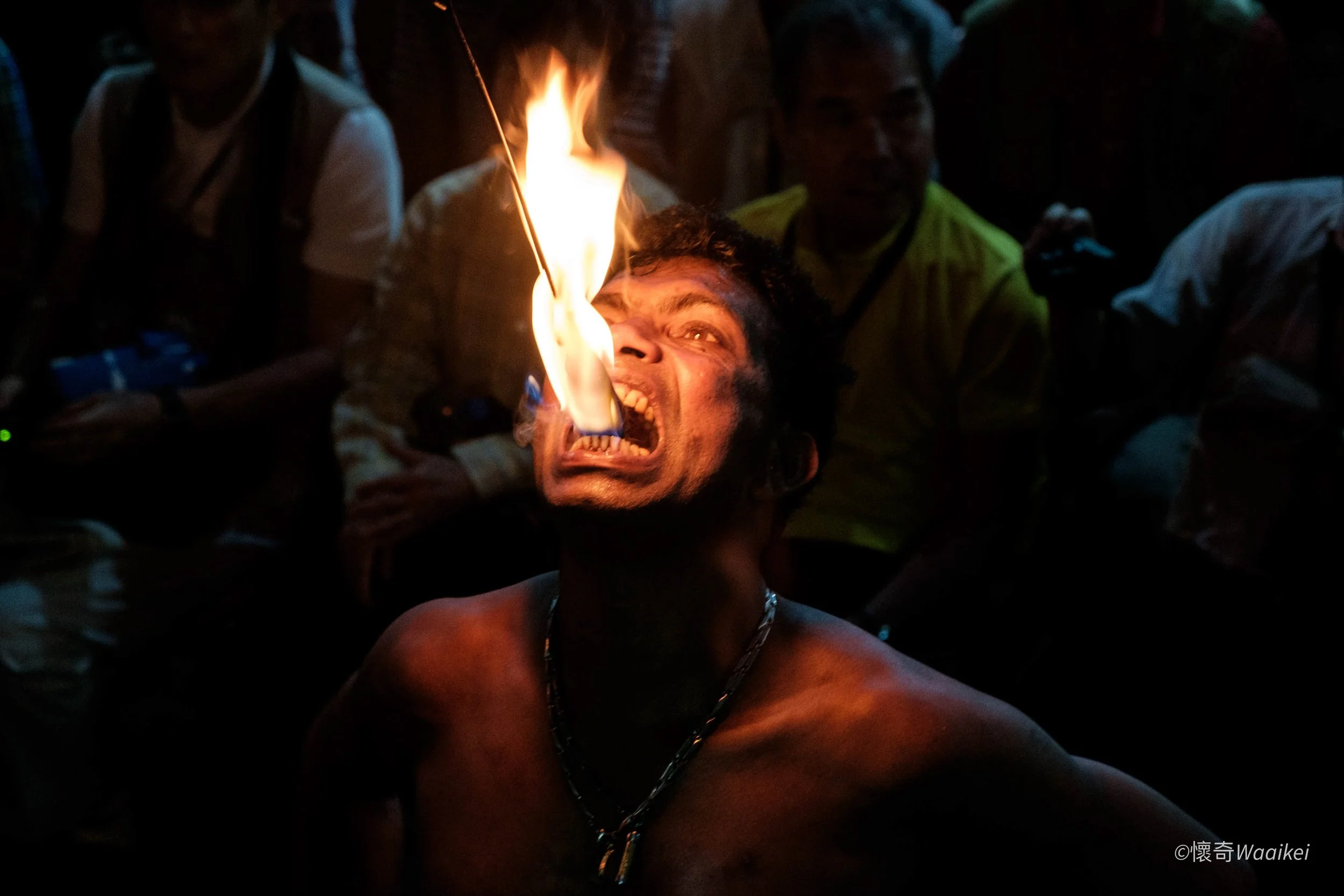 A person with curly hair is performing a fire-eating act, with fire emerging from their mouth, surrounded by a dark crowd.