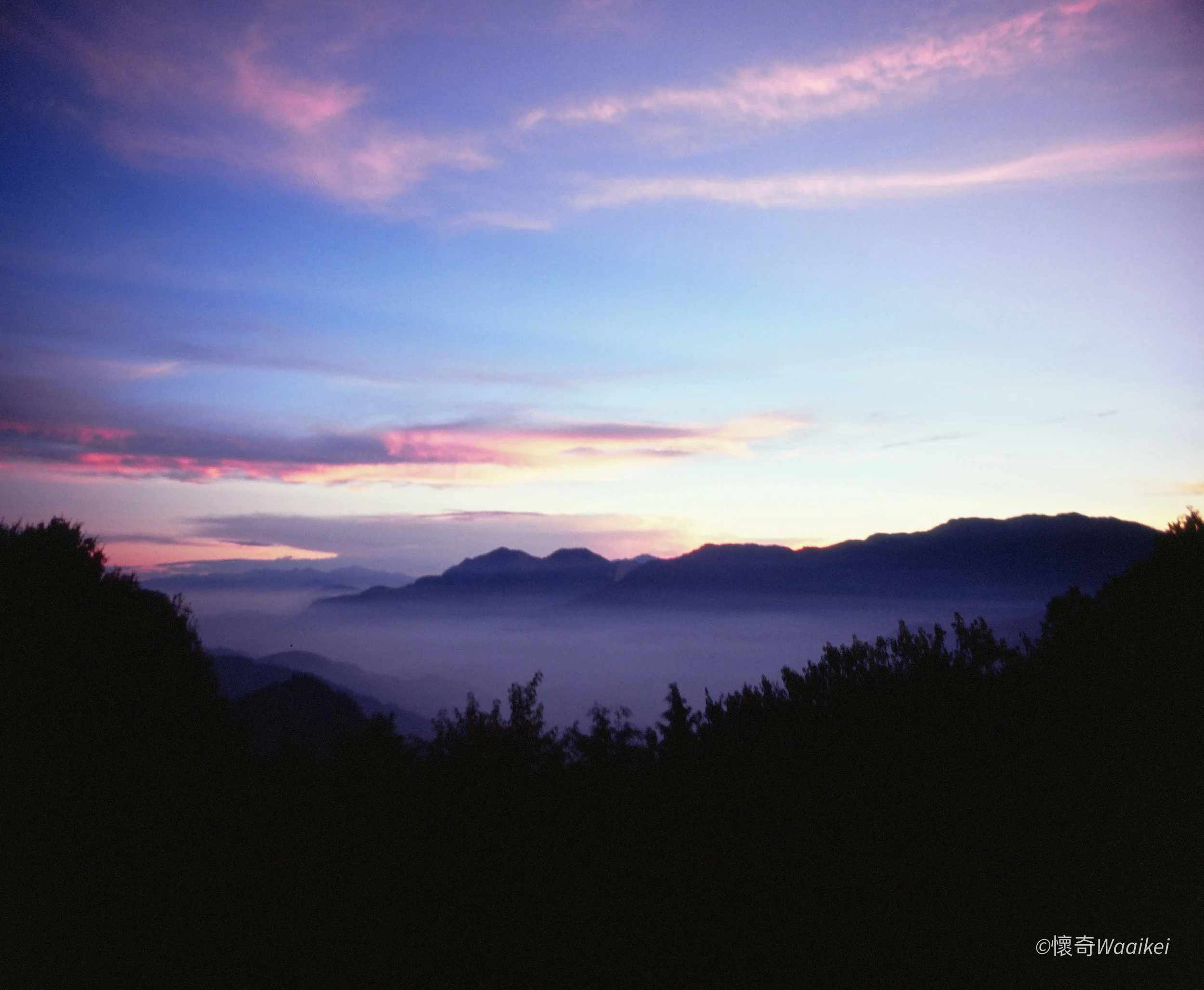 A scenic view of mountains at sunset with a sky filled with pink and purple clouds, and dark silhouetted trees in the foreground.