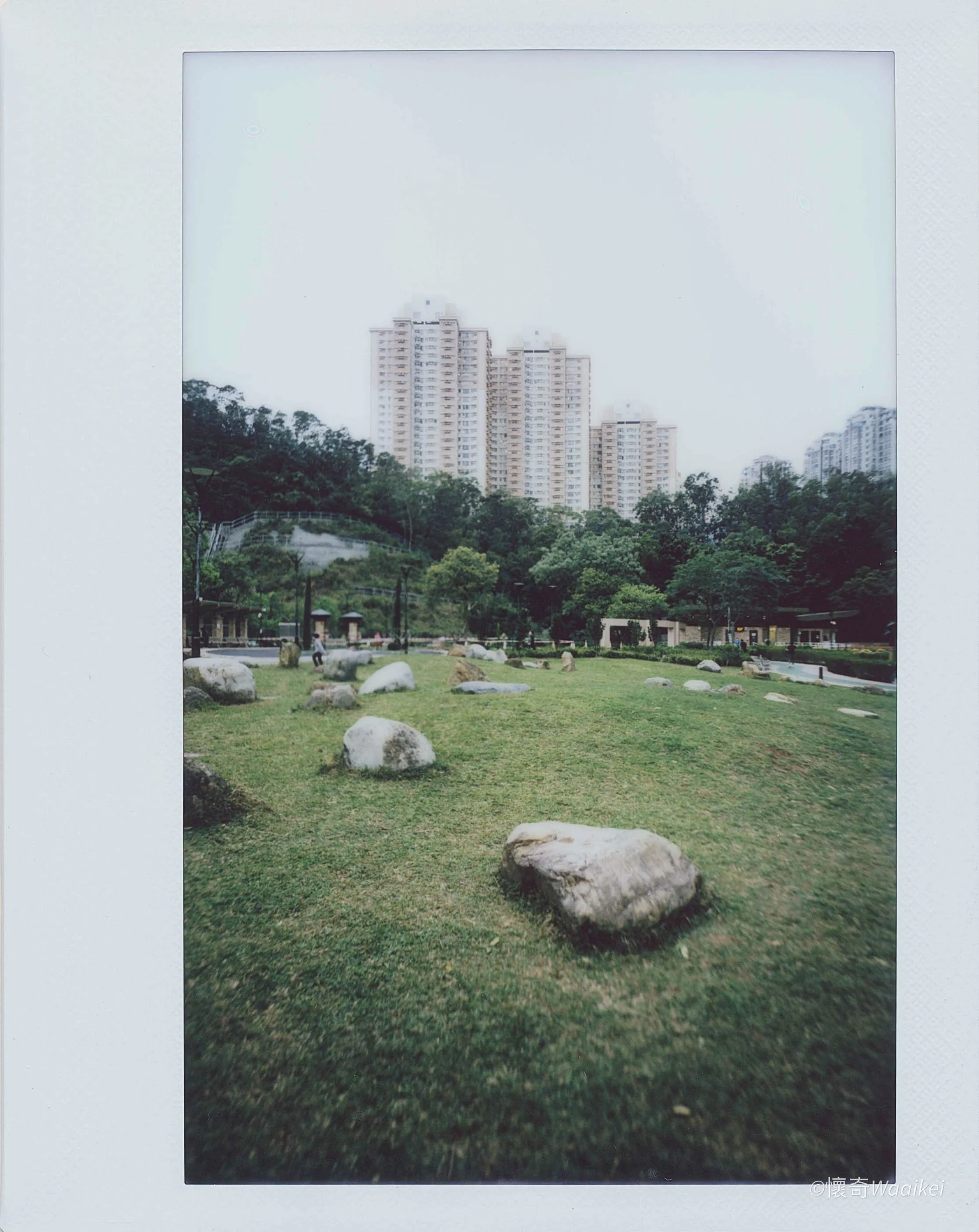City skyline with tall apartment buildings viewed from a park with rocks, trees, walking paths, and a playground.
