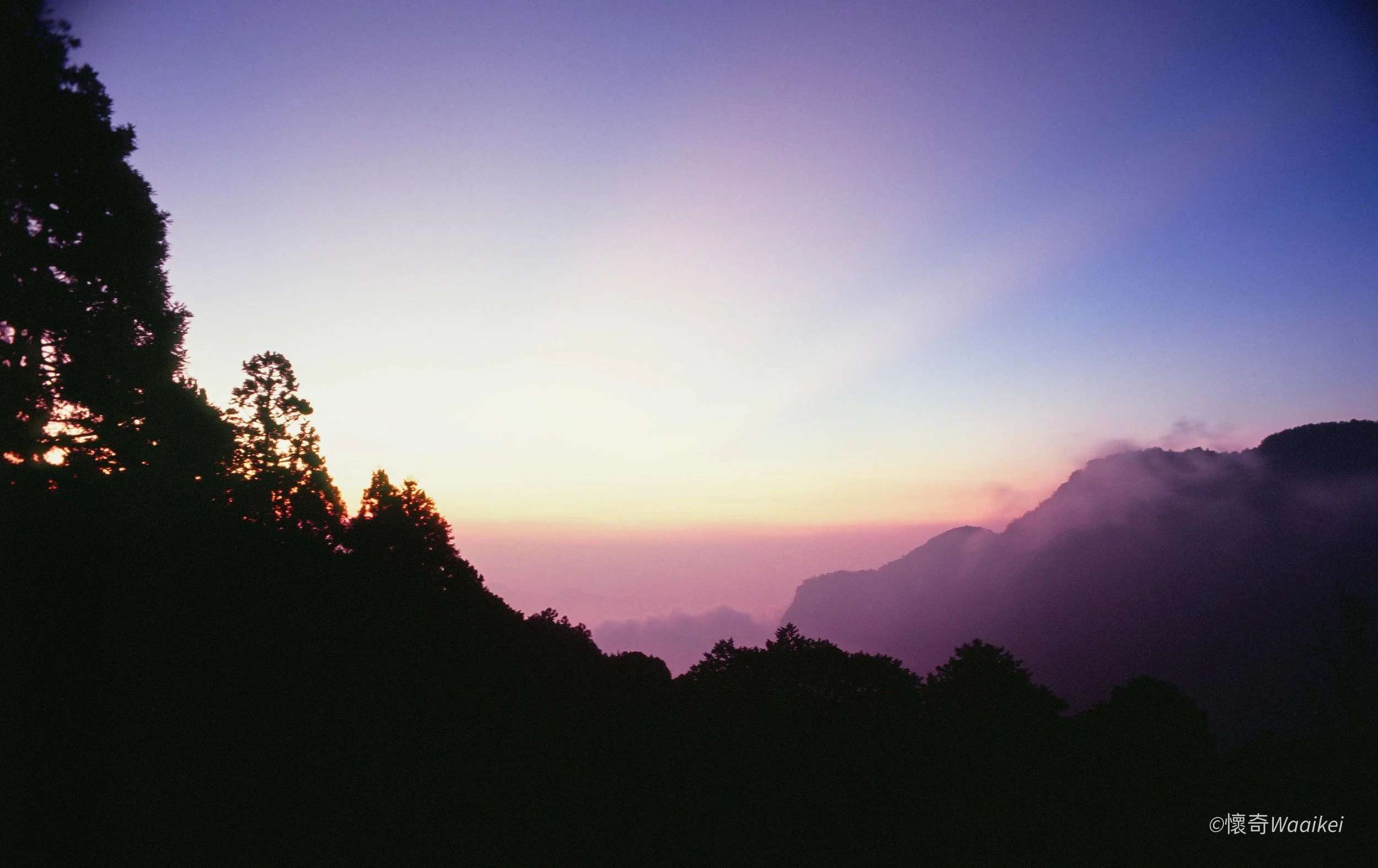 A mountain landscape during twilight with silhouettes of trees in the foreground and mountains in the background, sky fading from light to dark with hues of purple and pink.