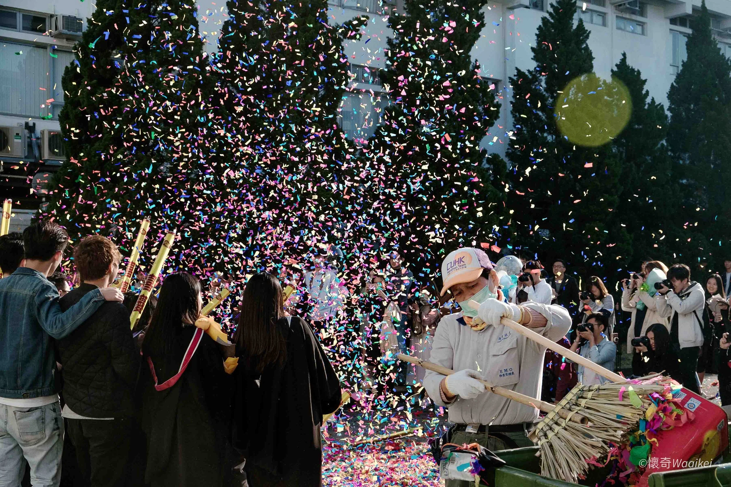 A crowd of people celebrating a festival outdoors, with a large Christmas tree decorated with colorful lights and ornaments. Confetti is falling from above, and some people are taking photos. One person appears to be holding a stream of paper firewor
