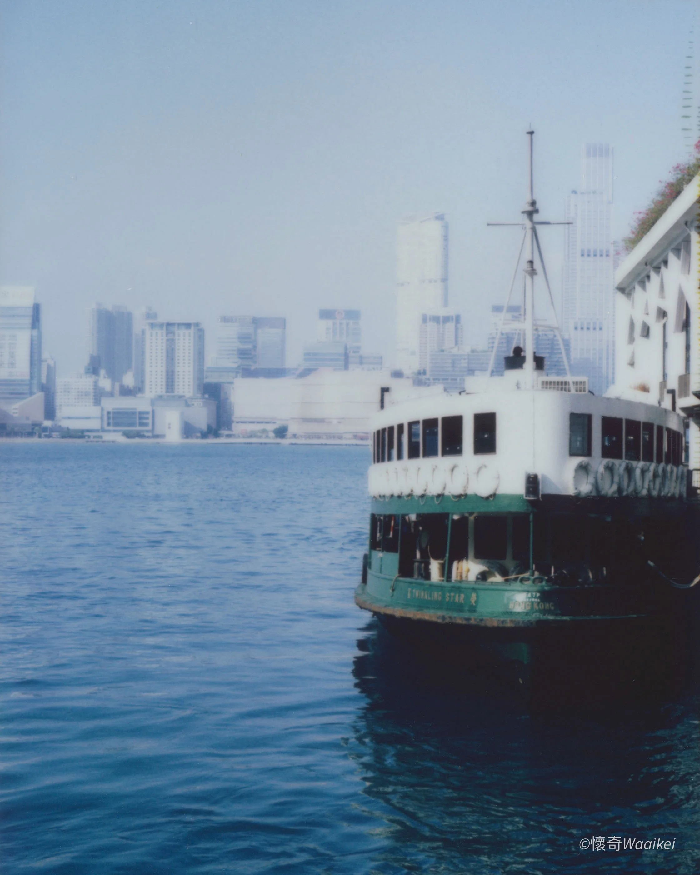 A ferry boat on the water with a city skyline in the background, including tall buildings and a cloudy sky.