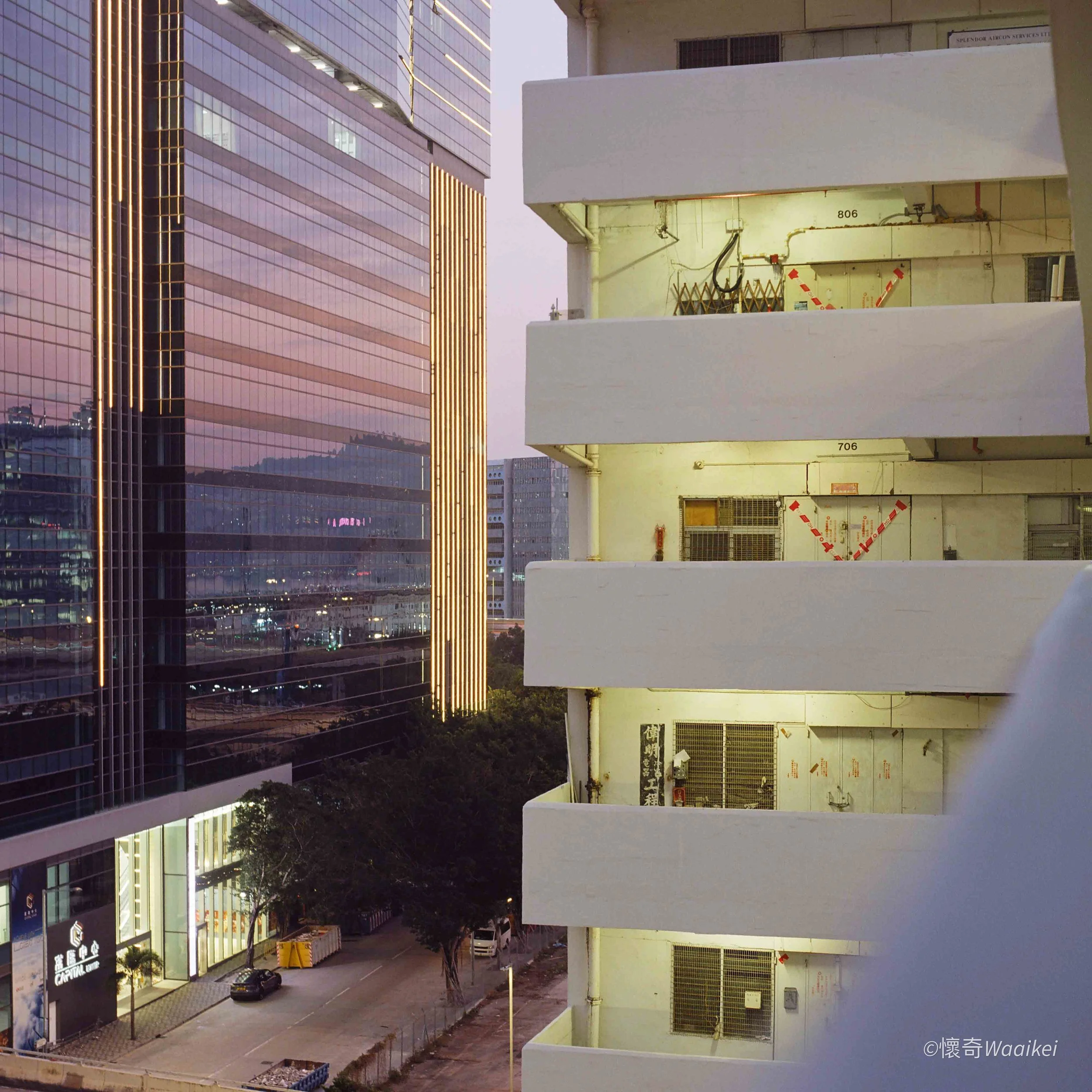 Cityscape at dusk showing a modern glass skyscraper with gold lighting accents on the left and a white building with multiple levels on the right, with a street and parked cars below.