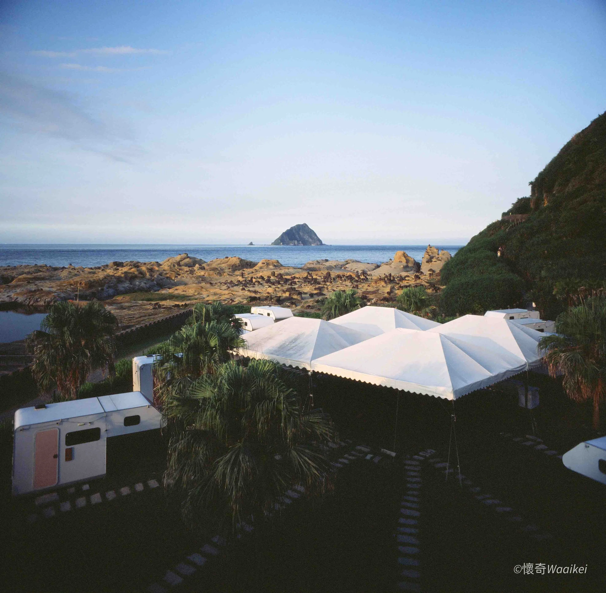A coastal scene at dusk with rocky shoreline, ocean, distant rock formation in the water, green hillside on the right, and white tents in the foreground.