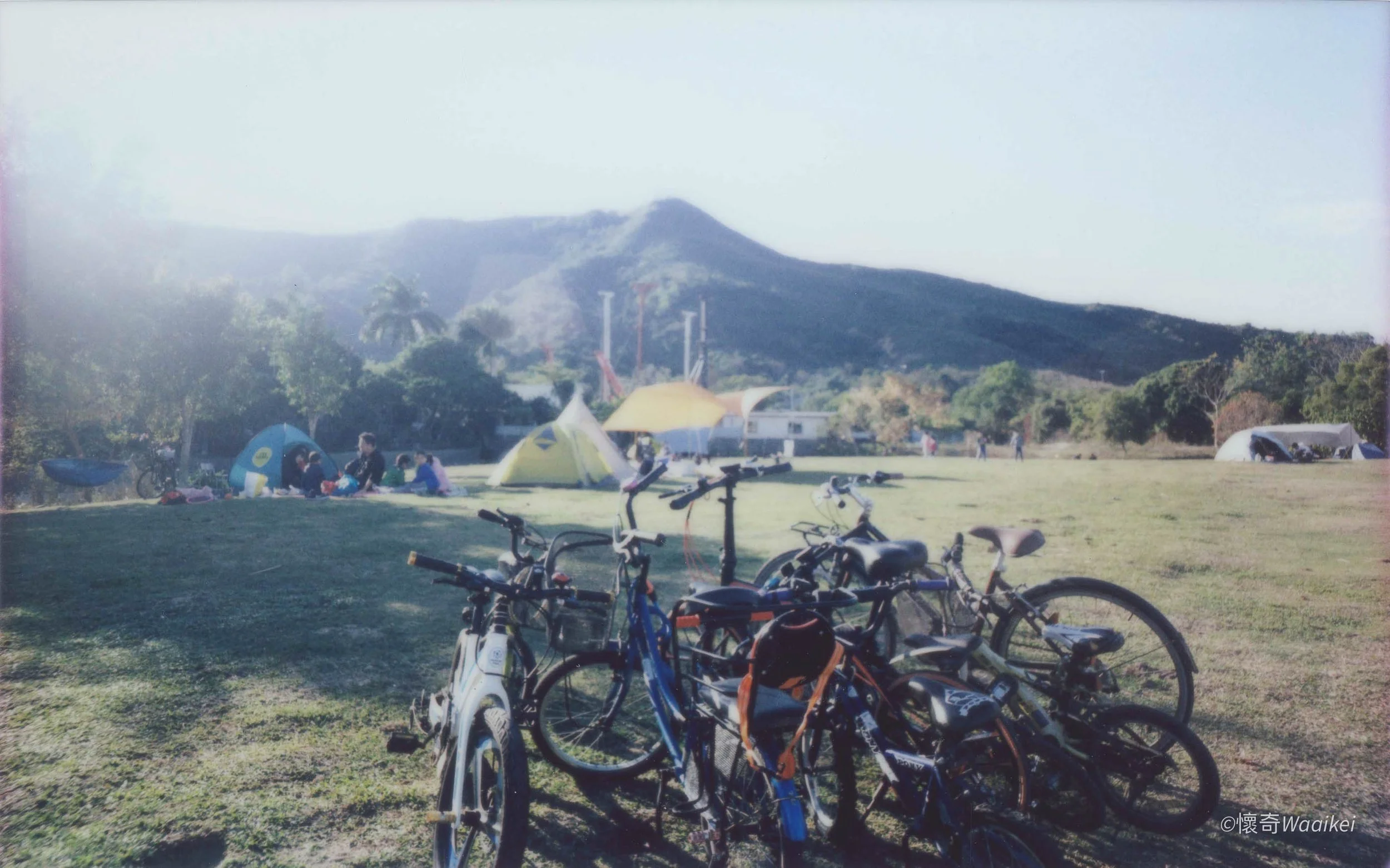 Bicycles parked on a grassy field with camping tents, and people sitting and standing nearby. In the background are trees and a mountain.