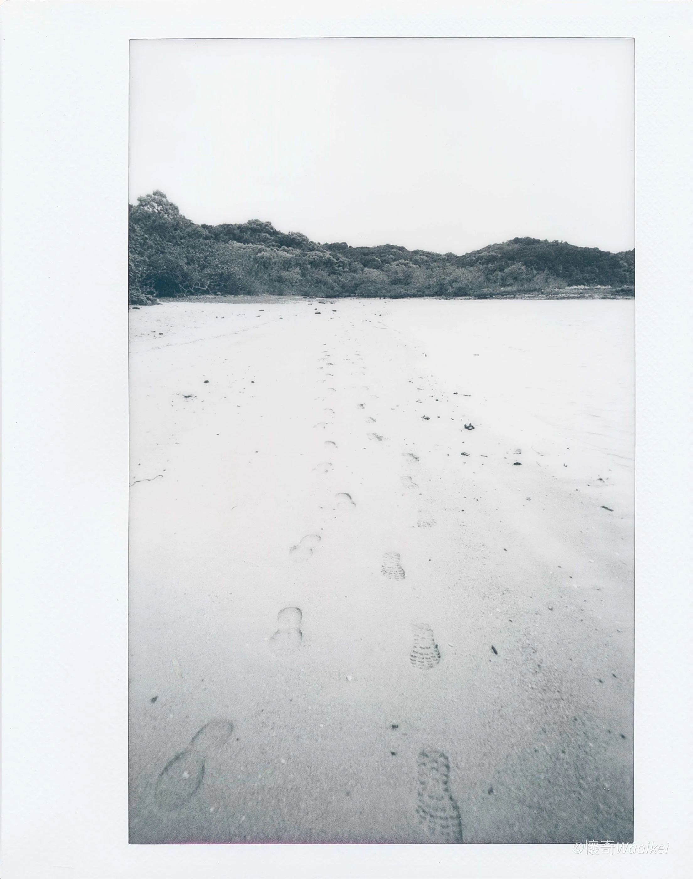 Black and white photograph of footprints in the sand along a shoreline, with trees and hills in the background.