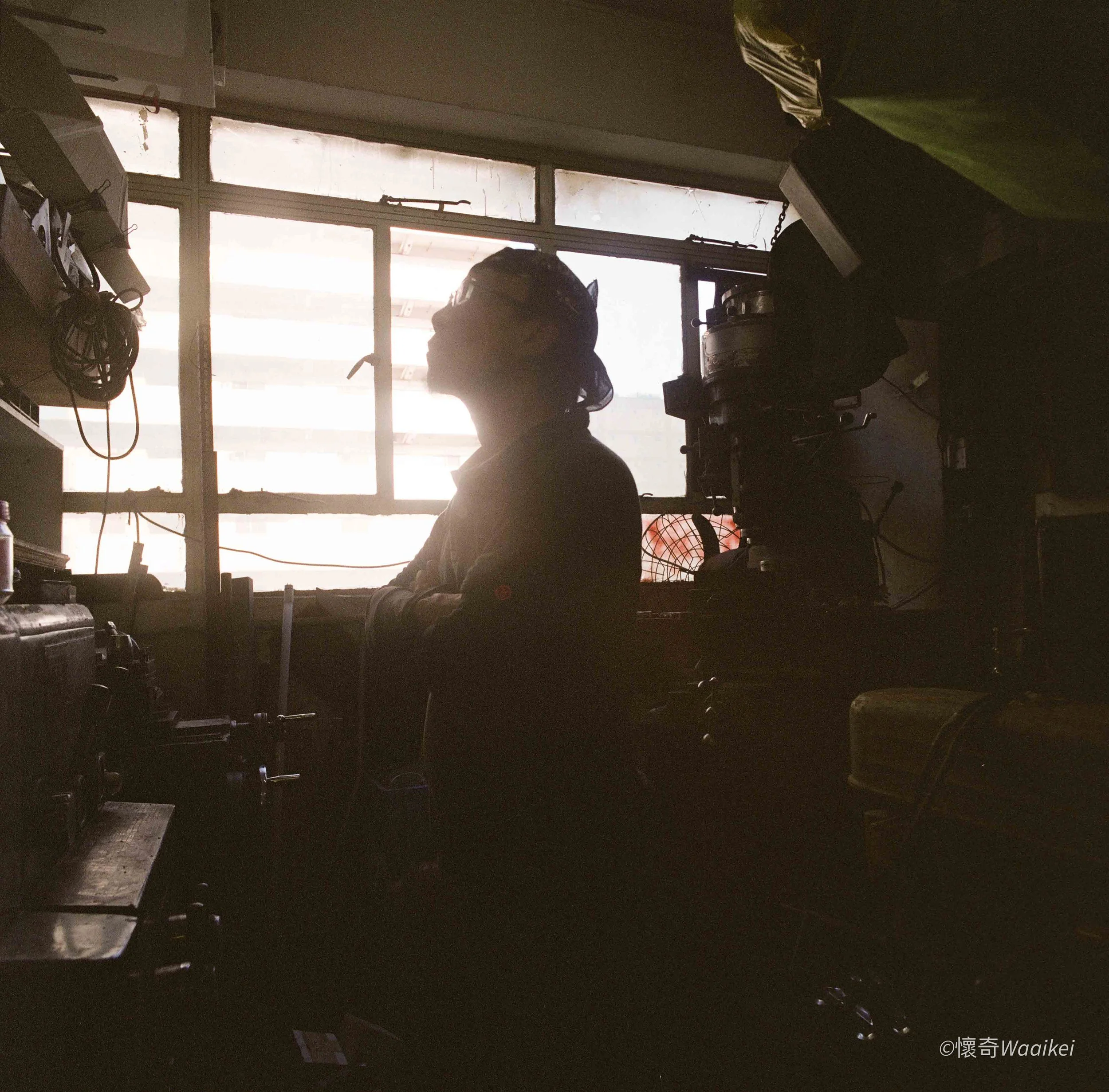 A person wearing a helmet working in a dimly lit room with a large window showing daylight outside. Various equipment and cables are visible around the person.