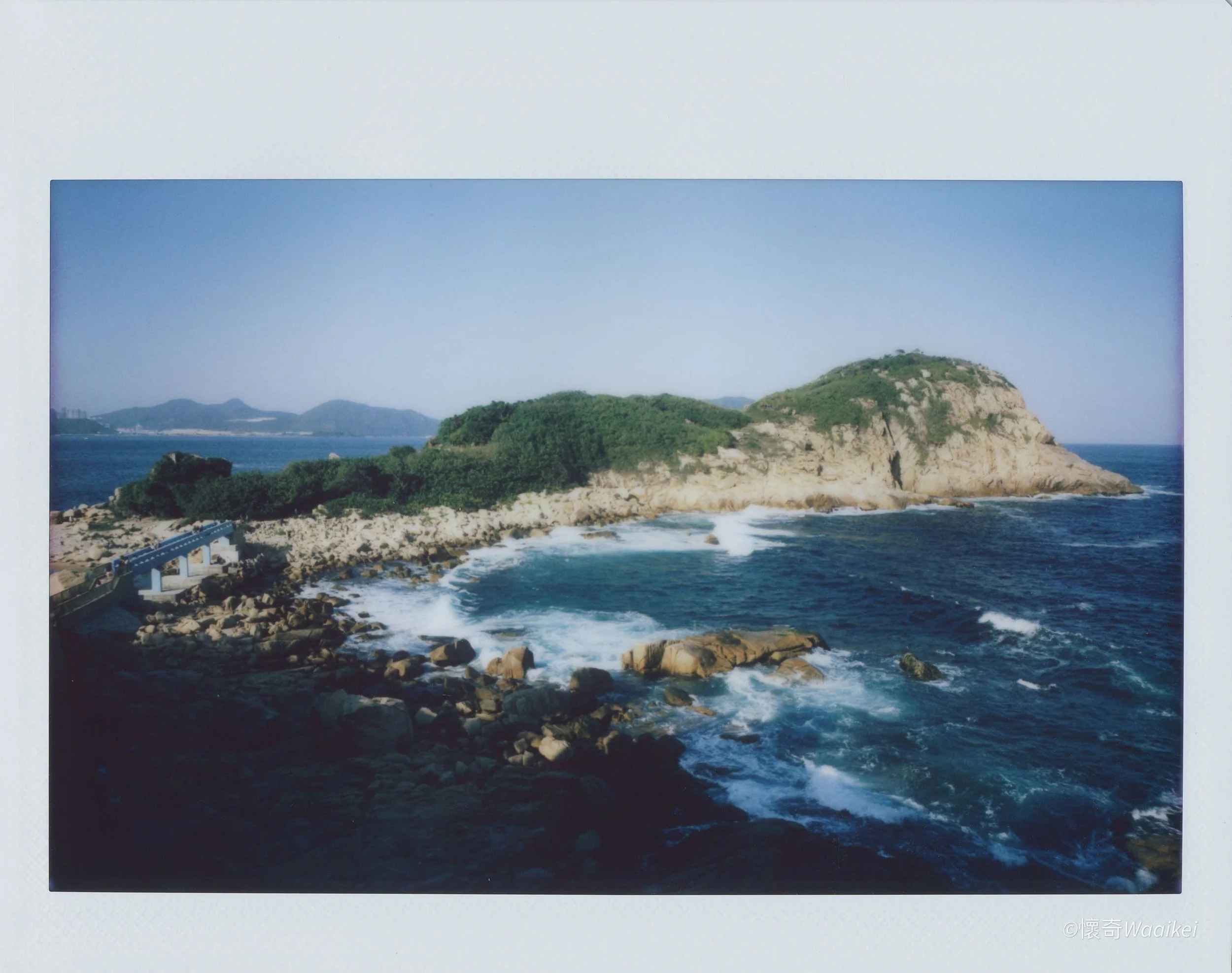 Photograph of a rocky coastline with waves crashing against rocks, a small hill covered with greenery, and a body of water with mountains in the background under a clear blue sky.