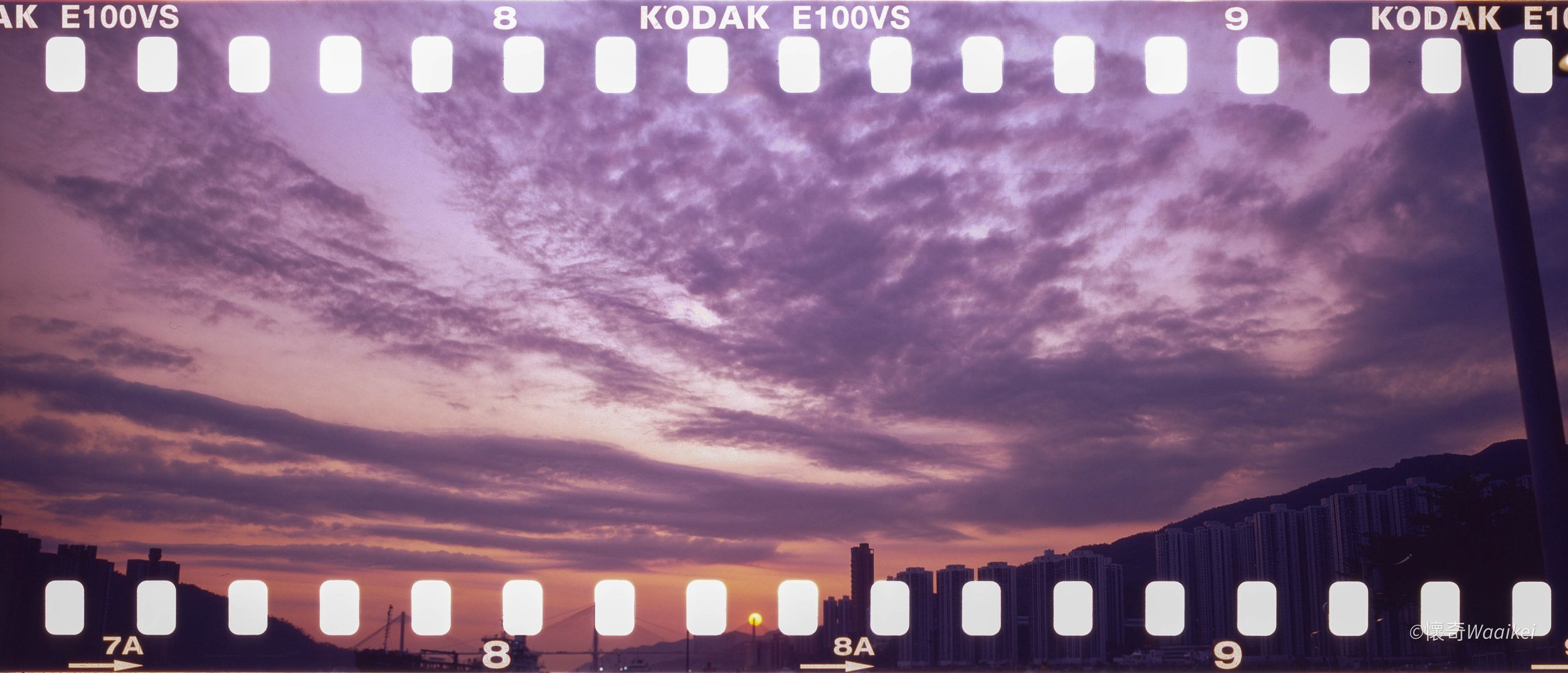 Sunset over a city skyline with purple and pink clouds, hills in the background, and a bridge silhouette in the foreground.
