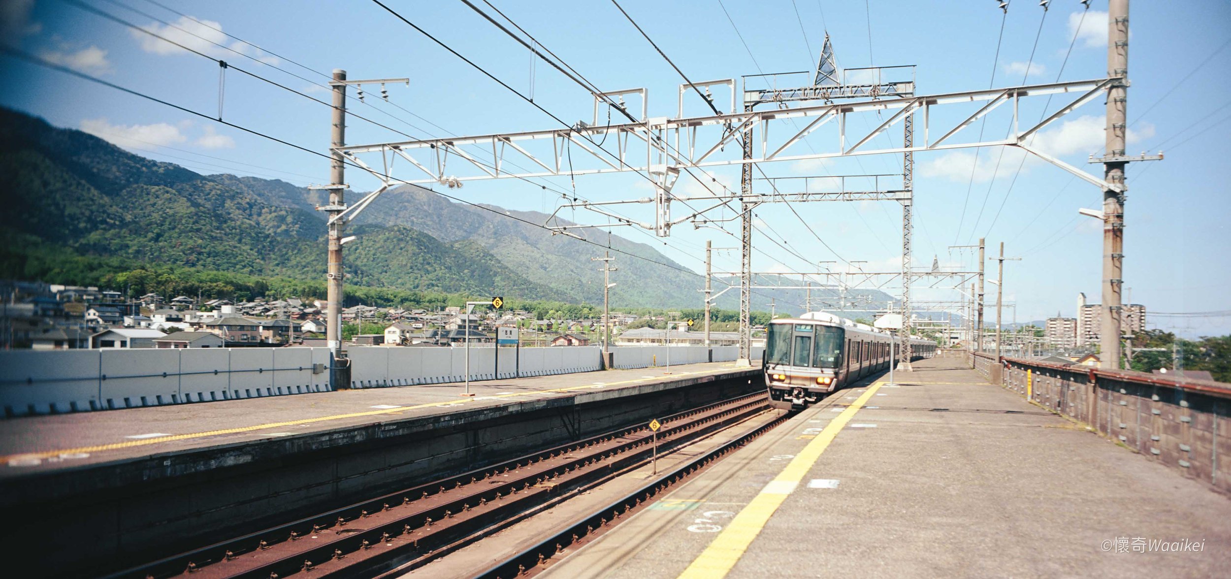 An electric train arriving at a train station platform with mountains in the background and a blue sky overhead.