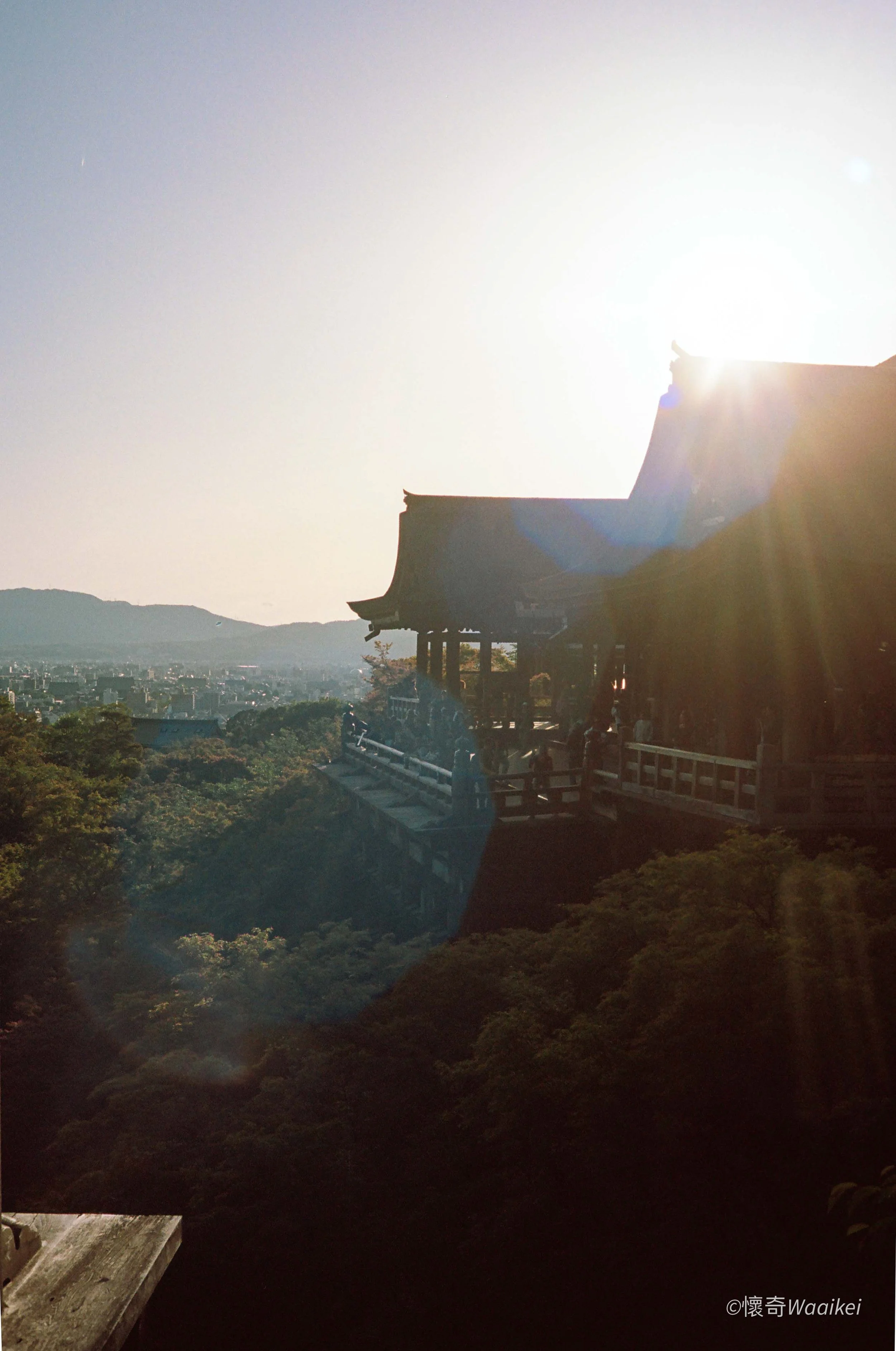 Sunset or sunrise over a traditional Japanese temple with a hilly landscape and lush green trees in the foreground.
