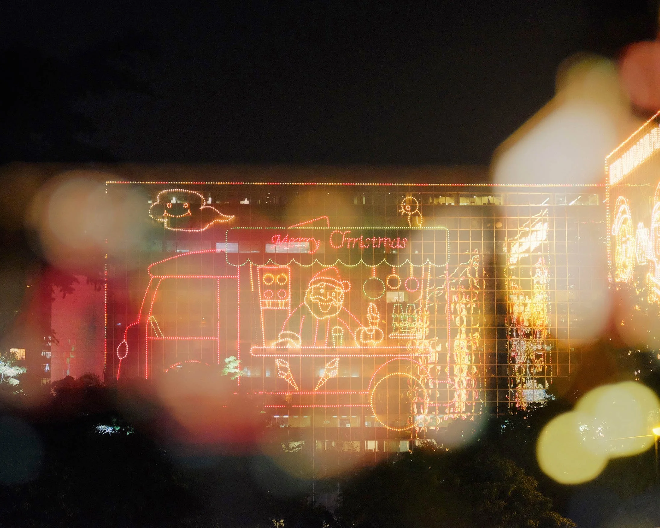 Nighttime scene of a building decorated with colorful Christmas lights shaped like Santa Claus, a reindeer, and other holiday figures, with a sign reading "Merry Christmas."