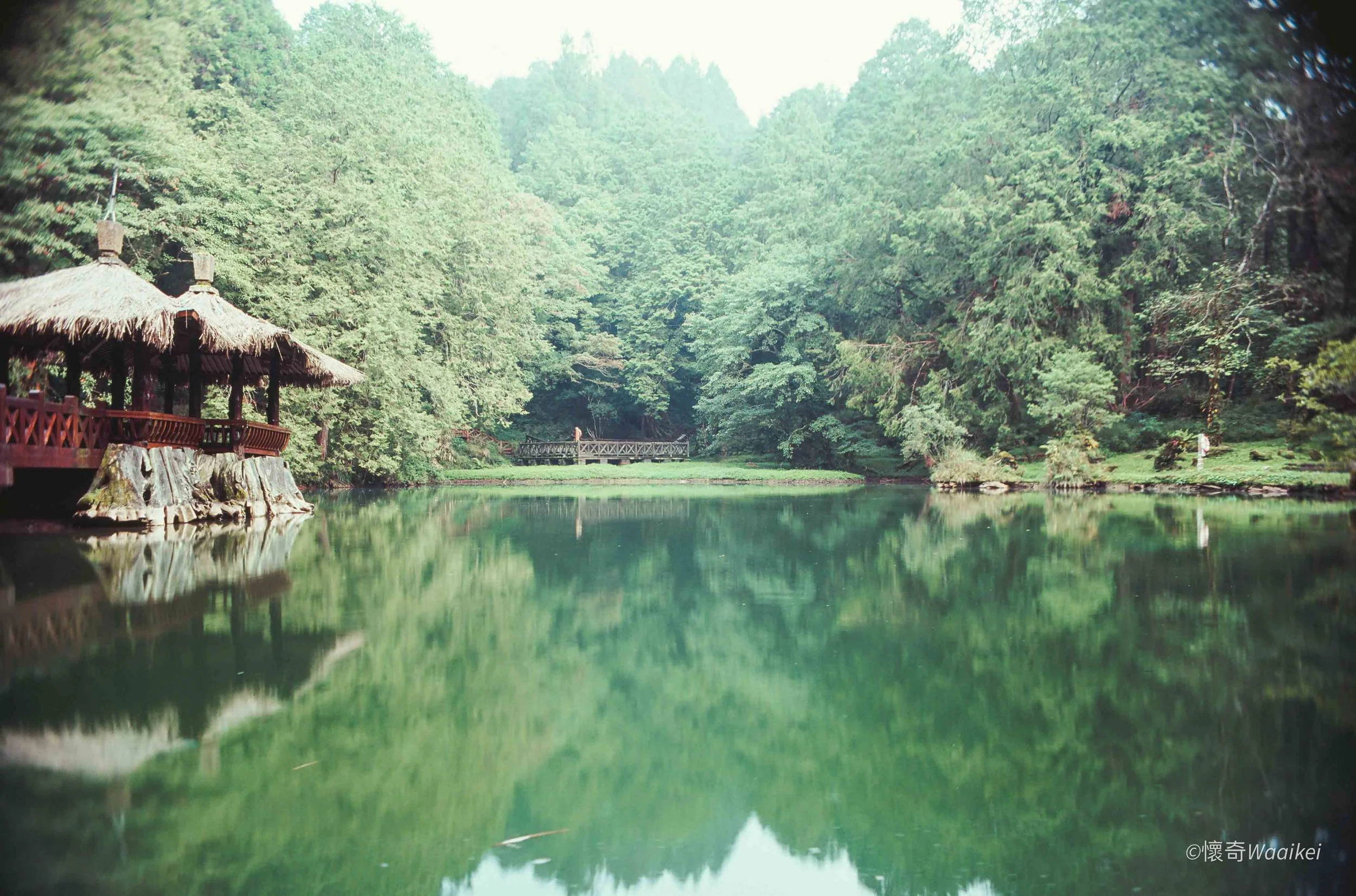 A peaceful pond surrounded by lush green trees with a wooden gazebo on the left and a small bridge in the distance.