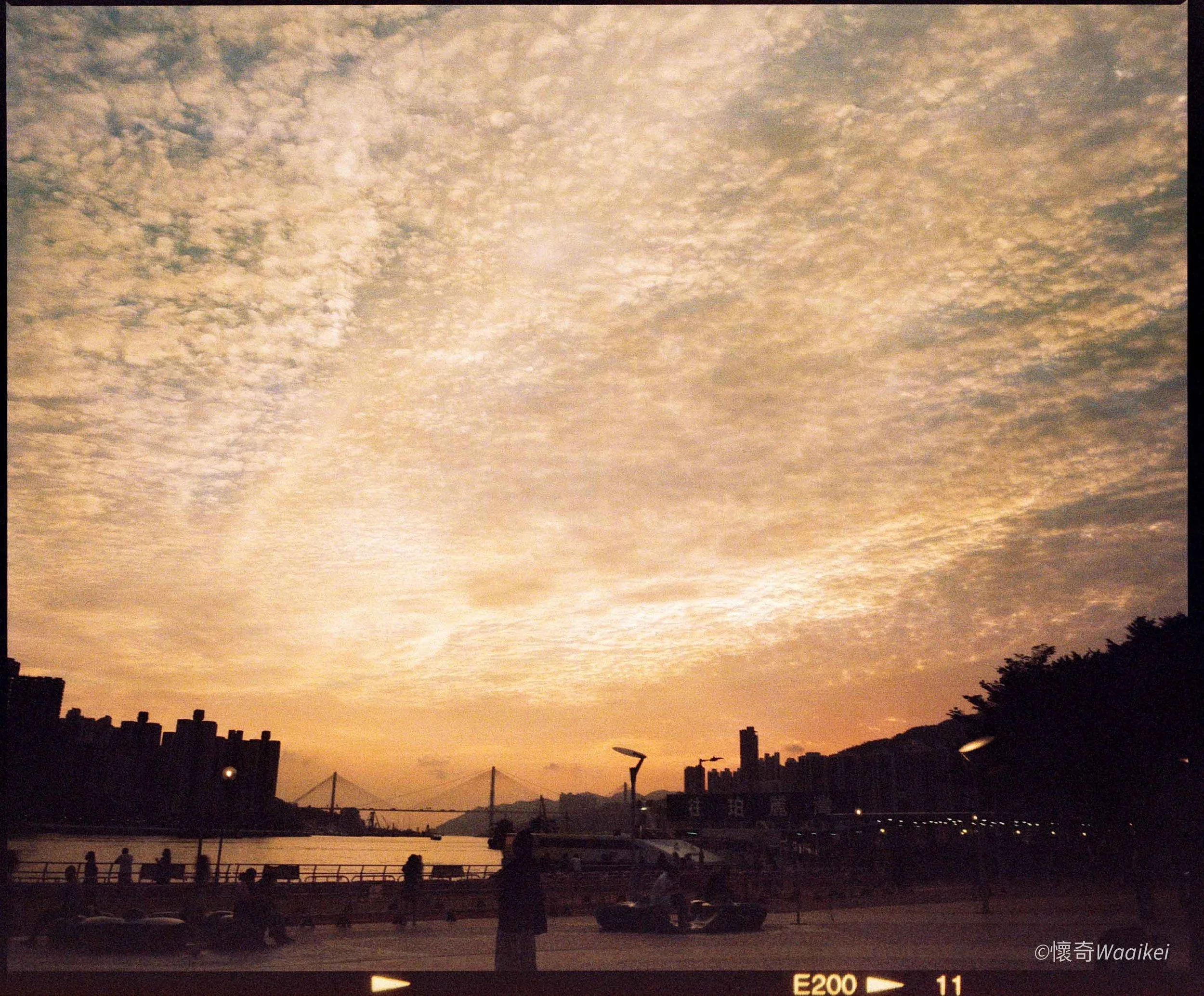 Sunset over a river with silhouettes of bridge, buildings, and people standing along the riverbank, under a sky with scattered clouds.