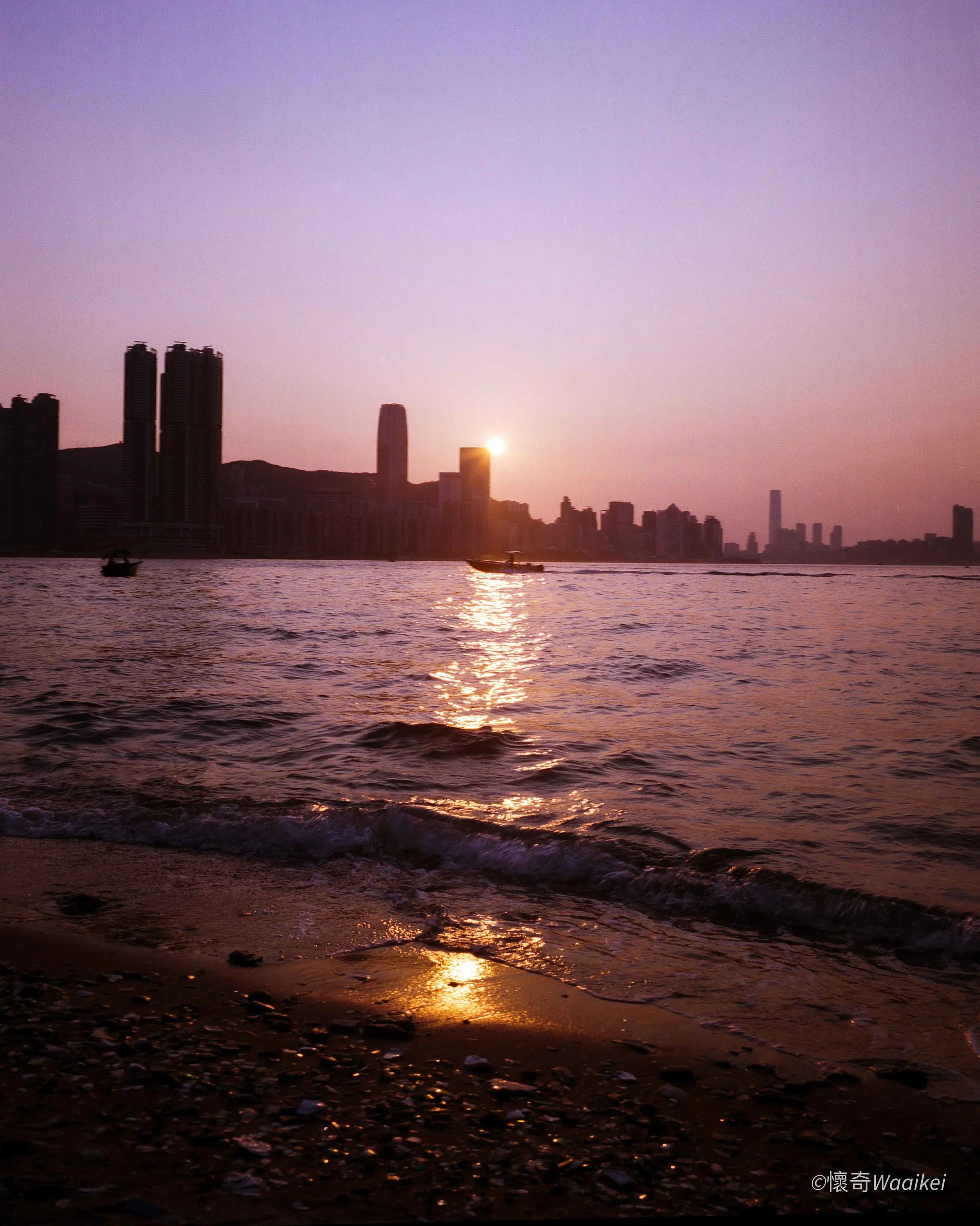 Sunset over a city skyline with high-rise buildings, with boats on the water and a boat on the shore, creating a warm glow reflecting on the water.