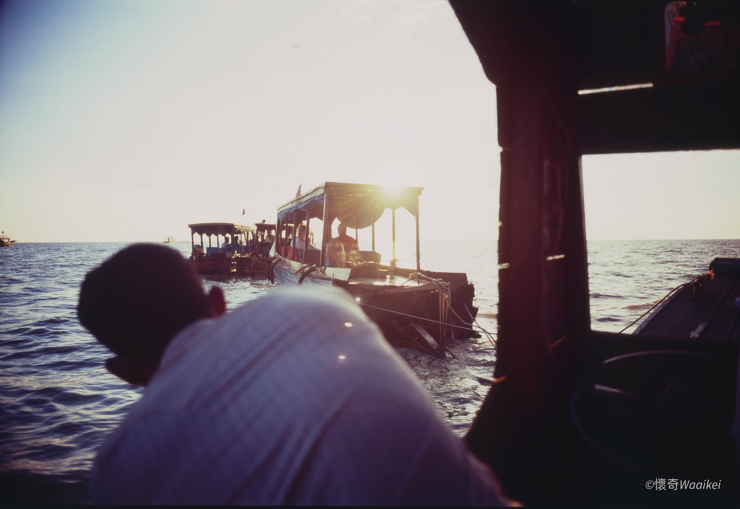 View of boats on the water during sunset, with part of a dock or structure in the foreground, creating a silhouette against the bright sky.