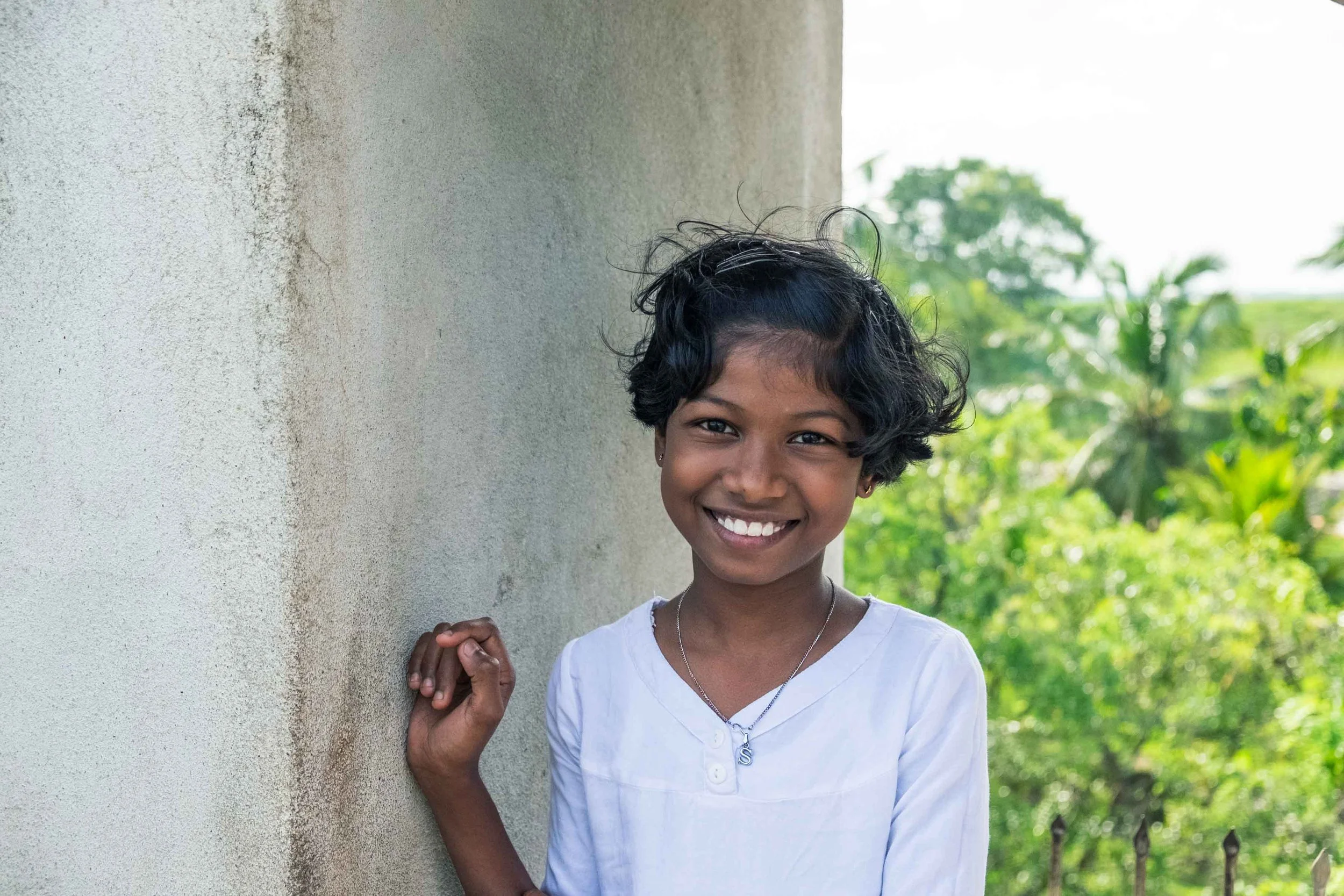 A young girl with short curly black hair, smiling, wearing a white shirt and a necklace with an S-shaped pendant, standing outdoors near a textured wall with greenery and trees in the background.