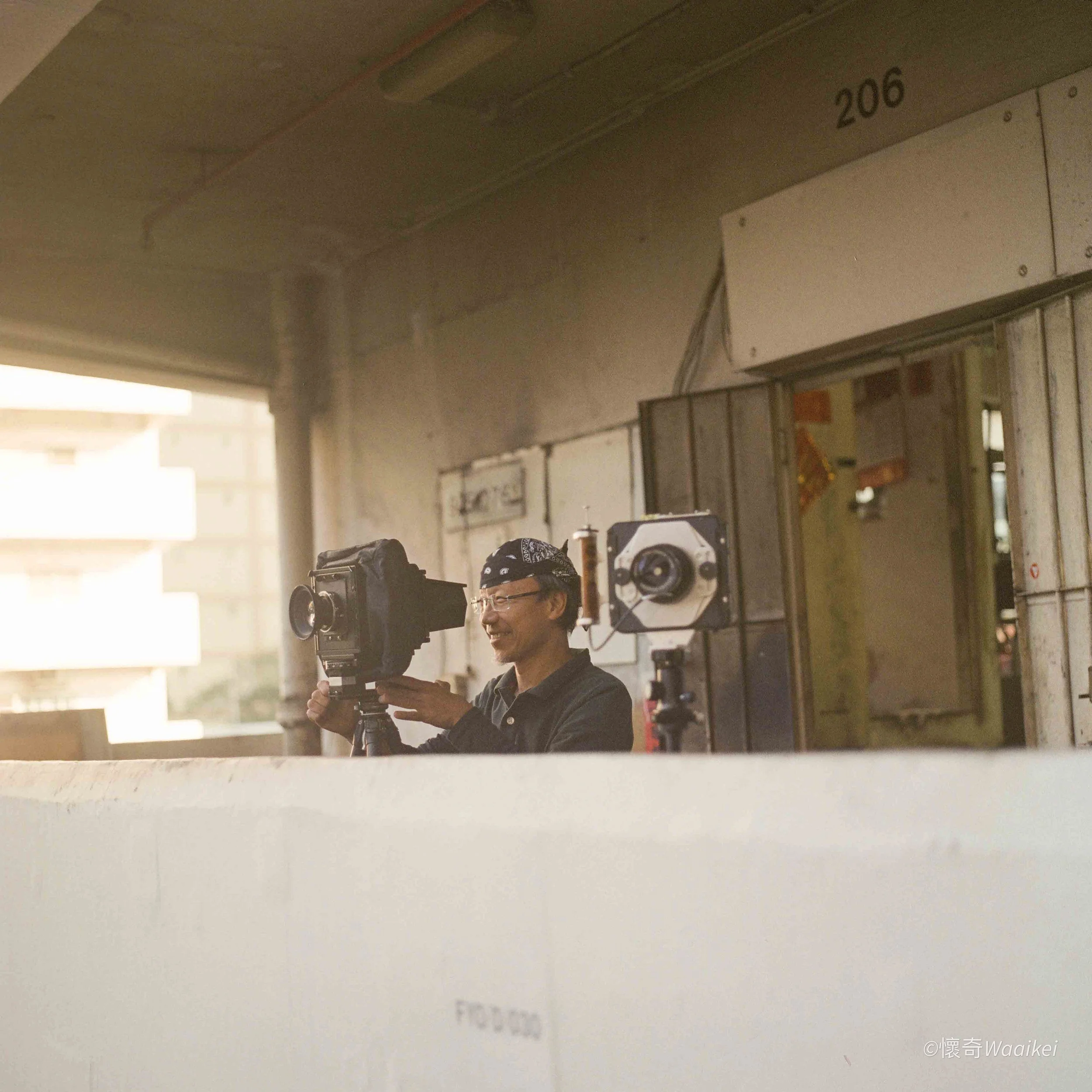 The image shows a man wearing glasses, a black bandana, and a dark jacket operating a camera on a tripod in a room with air conditioning units and electrical panels on the wall. There is a white wall with some colorful posters or notices behind him. 