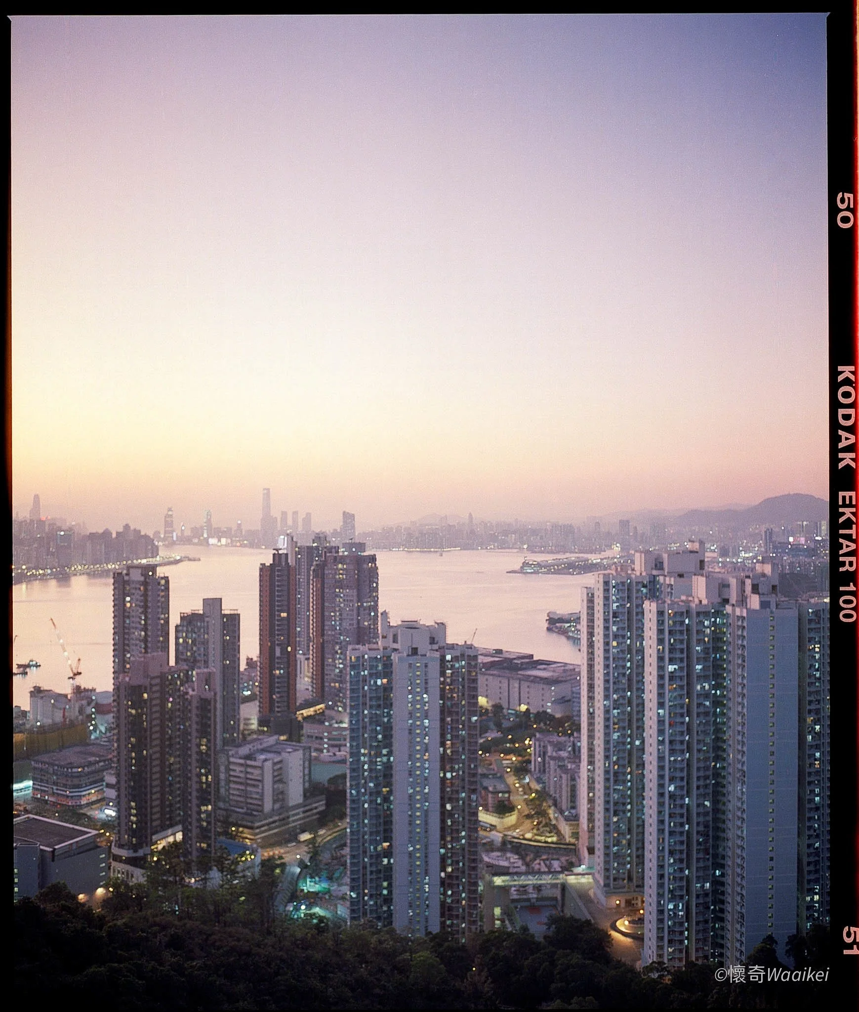 View of a city skyline at dusk with high-rise buildings, a river, and a mountain range in the background, under a hazy sky.