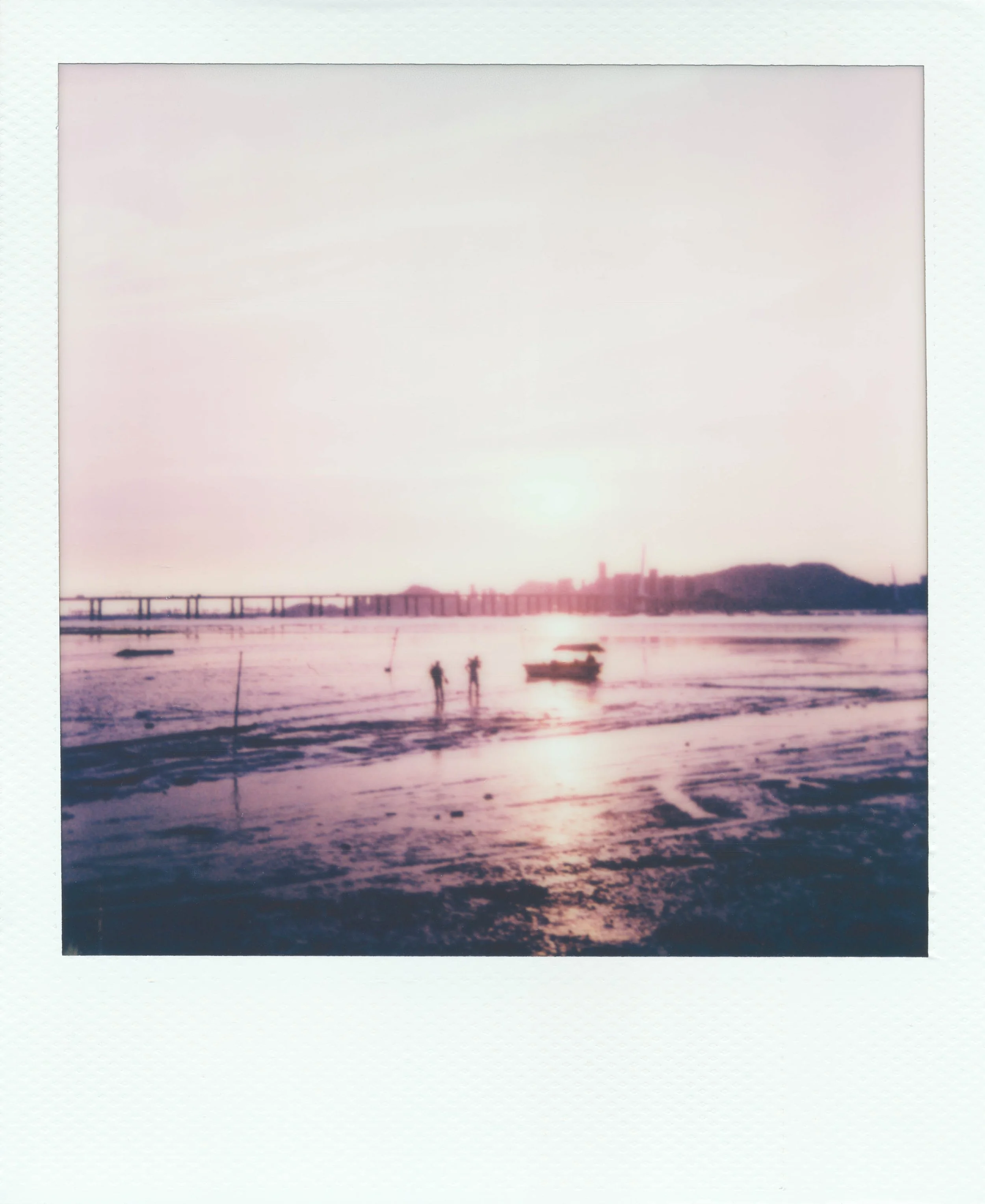 Polaroid photo of a sunset over a river with a bridge, two people walking, and a boat in the water.