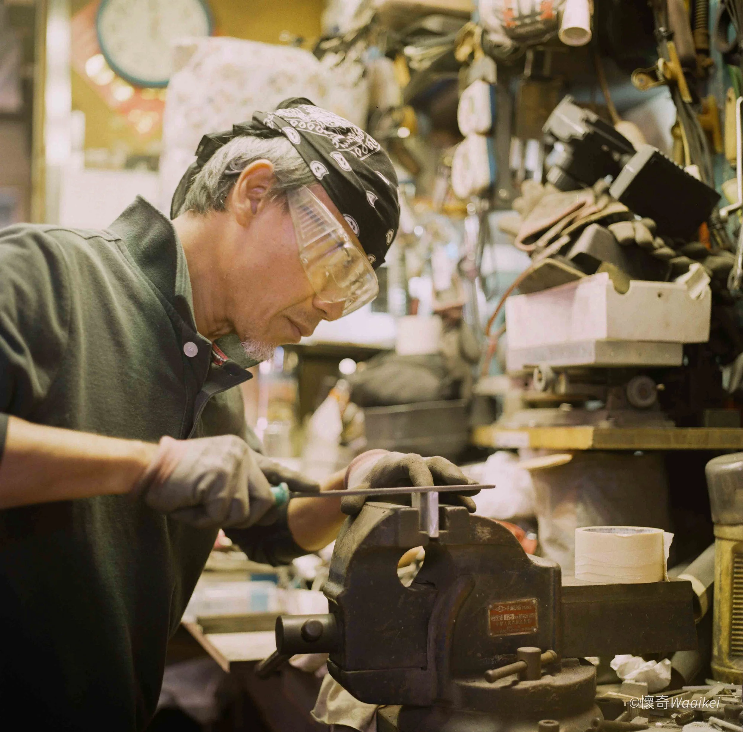 A man working on a machine at a workshop, wearing safety glasses, a bandana, and gloves, with various tools and equipment in the background.