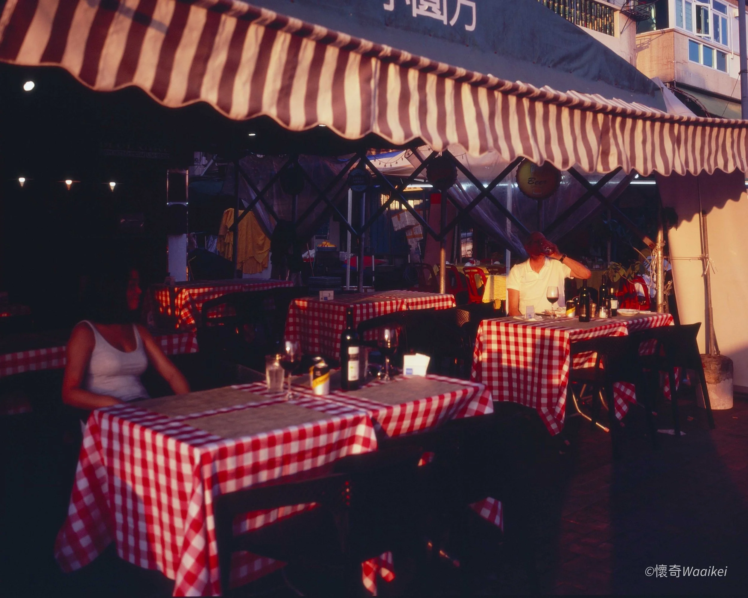 Outdoor restaurant area with tables covered in red and white checkered tablecloths, under a striped awning, during evening with warm lighting, a person sitting at a table drinking from a glass, with bottles on the table.
