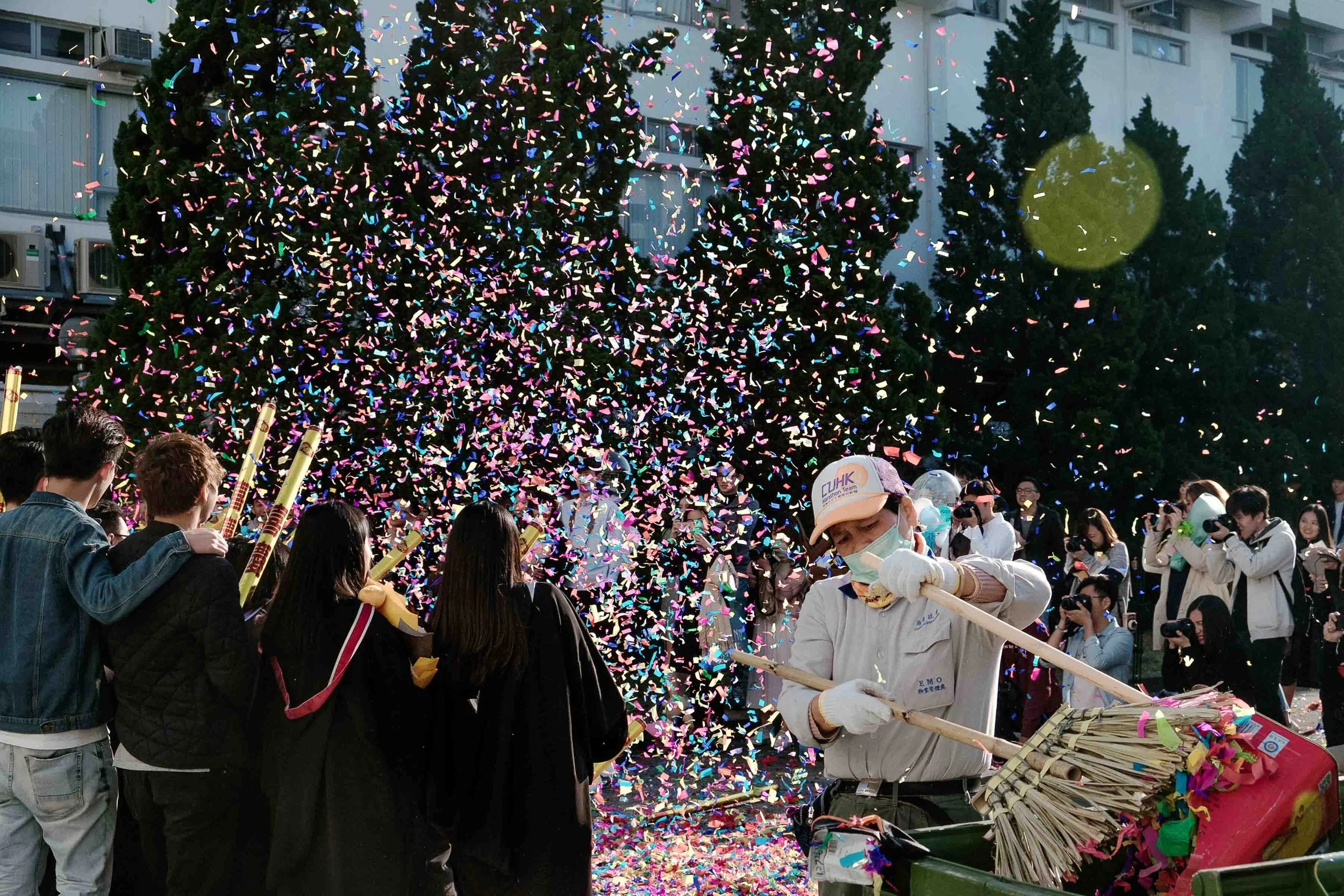 People gather outdoors during a celebration with colorful confetti falling from above. There is a mix of men and women, some taking photos, and a person in a white jacket with gloves handling a confetti launcher. In the background, there are trees an