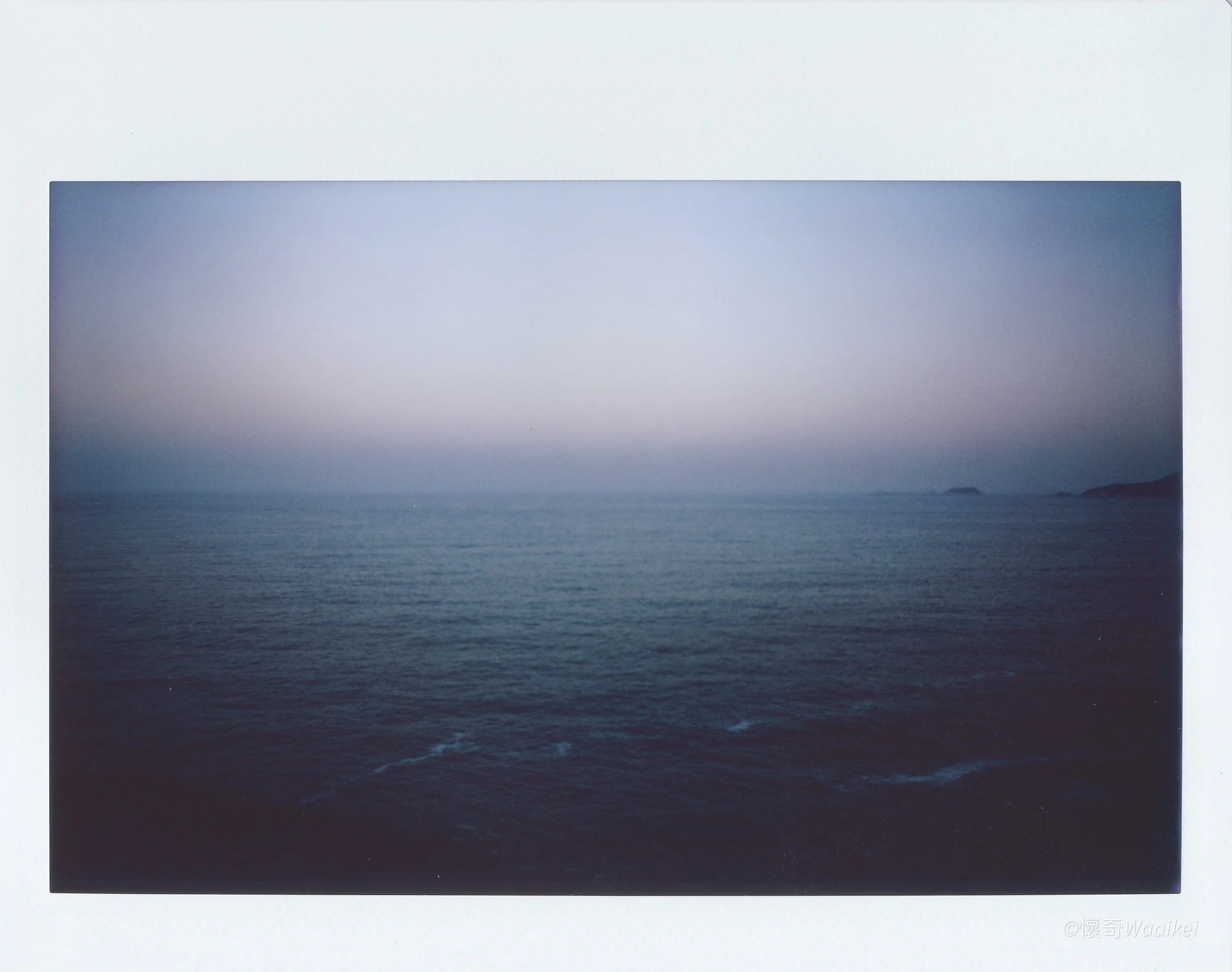 A Polaroid photo of the ocean with a distant island, taken during dusk or dawn, with a gradient sky from light to darker hues.