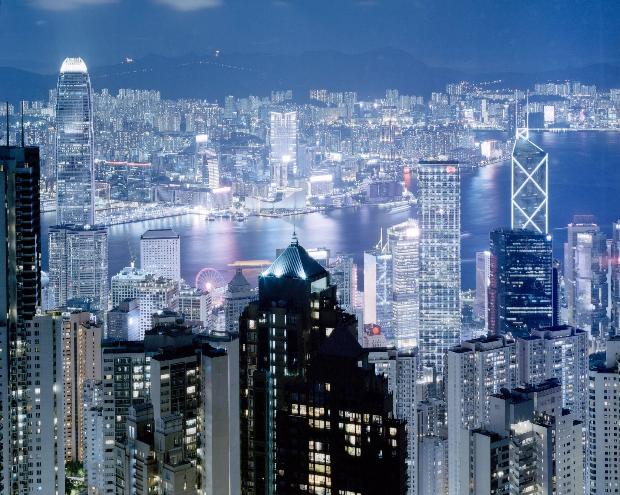 Nighttime view of a city skyline with illuminated skyscrapers and a body of water reflecting city lights, featuring tall buildings with bright lights and mountain silhouettes in the distant background.