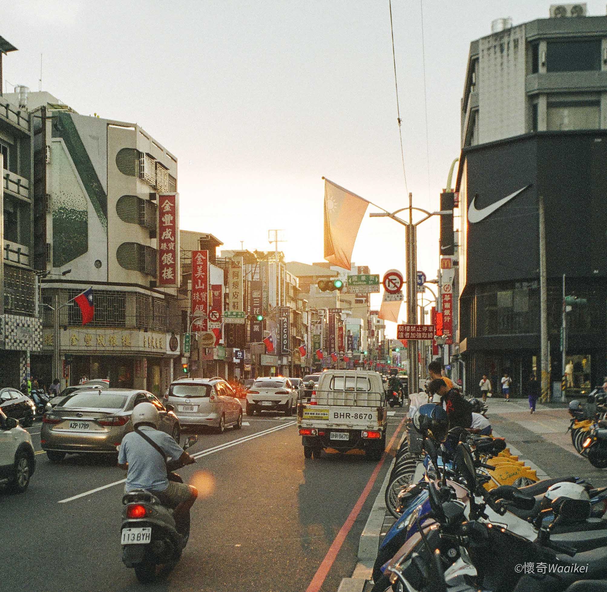 A busy city street with cars, motorcycles, and bicycles parked along the curb. Buildings with storefronts and signs with Chinese characters are visible. The sky is overcast with the sun setting, and traffic lights are green. People are walking on the